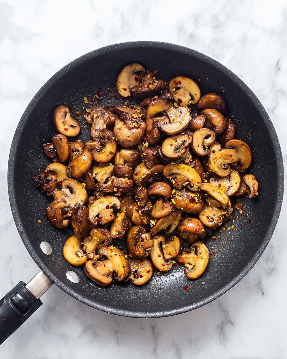 A black frying pan filled with cooked sliced mushrooms, showing a mix of light and dark brown colors with a slightly shiny texture from cooking. The mushrooms vary in size and shape, scattered around the pan, with some browned edges. Small yellow and red spices are sprinkled unevenly on top, adding contrast to the mushrooms. The pan is set on a white marbled surface, and the pan handle is visible at the bottom left corner. photo taken with an iphone --ar 4:5 --v 7