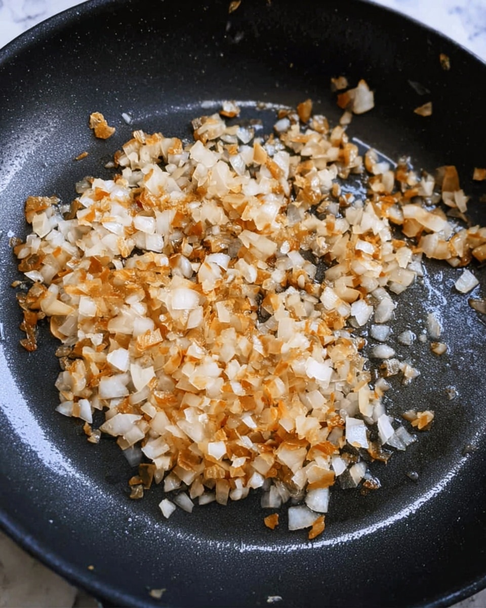 The image shows a close-up of a black frying pan with one layer of small, chopped onions being cooked. The onions are golden brown and white, with some pieces slightly darker from frying, giving a mix of soft and crispy texture. The pan has a shiny surface with light reflections around the onions. The background is a white marbled texture. photo taken with an iphone --ar 4:5 --v 7