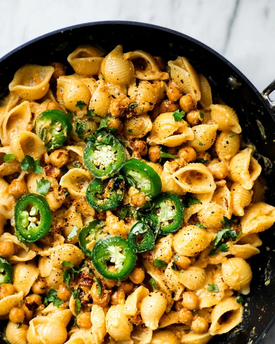 A close-up view of one-layer small shell pasta cooked creamy with a light orange sauce, mixed with small browned chickpeas spread evenly throughout. On top, several bright green sliced jalapeño peppers add a sharp contrast in color and texture. There are scattered fresh cilantro leaves adding small pops of green. The dish is in a black pan, sitting on a white marbled surface. The pasta and sauce have a smooth, slightly glossy texture with visible black pepper specks sprinkled all over photo taken with an iphone --ar 4:5 --v 7