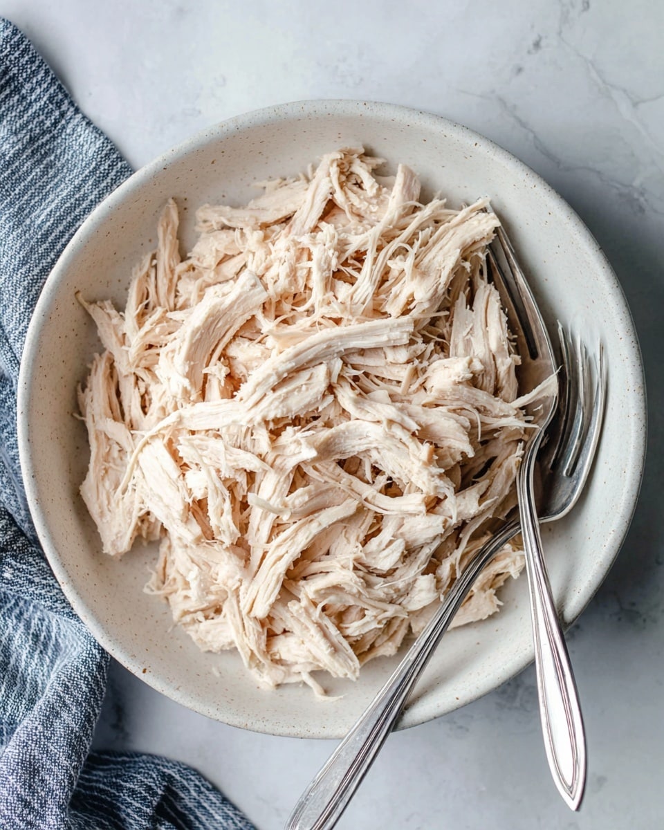 A white bowl filled with shredded pale beige chicken, showing thin and fibrous strands layered loosely across the bowl. Two silver forks lie on the right side inside the bowl, resting on top of some shredded chicken. The bowl sits on a white marbled surface with subtle grey veins, and a blue and white striped cloth is partly visible on the lower left side. The photo taken with an iphone --ar 4:5 --v 7