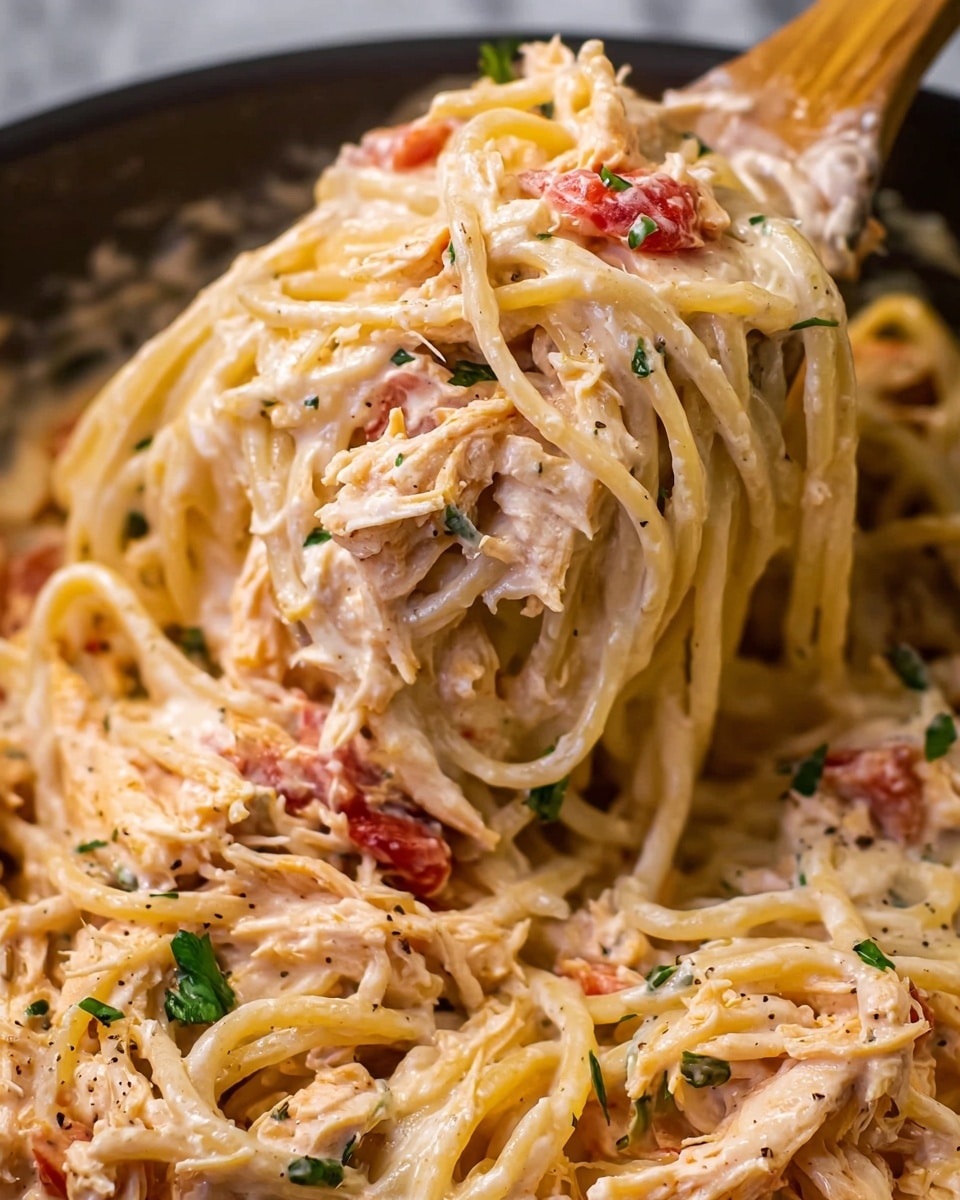 This close-up image shows creamy spaghetti pasta mixed with shredded pieces of light pink chicken and small chunks of red tomato. The pasta is coated in a thick, smooth, off-white sauce with a slight orange tint and specks of black pepper. Scattered green herb leaves add a fresh touch throughout the dish. The pasta is being lifted by a wooden spoon from a large serving bowl. The background is a white marbled texture. photo taken with an iphone --ar 4:5 --v 7