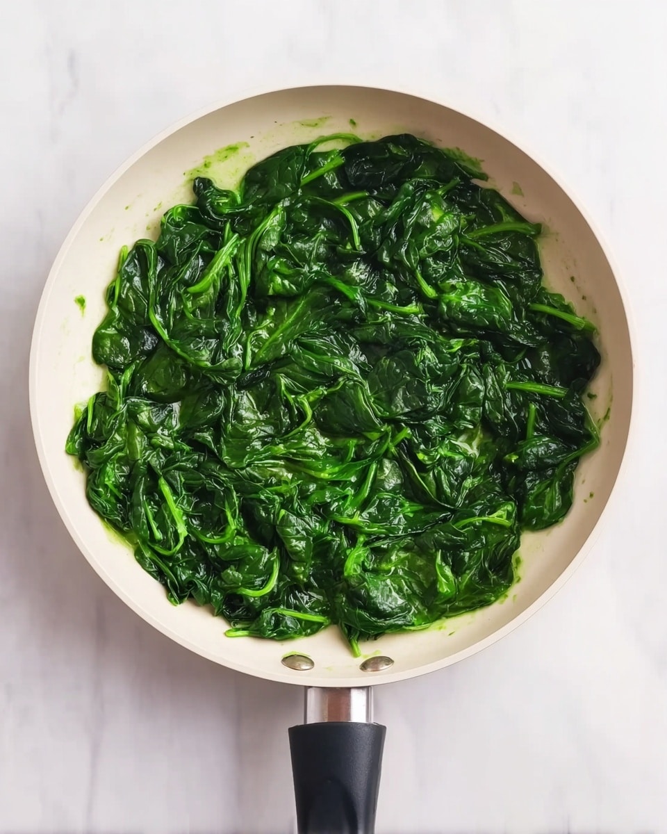 A white pan filled with cooked spinach leaves that are dark green and soft, evenly spread across the pan surface. The spinach looks slightly wet with a light shine, showing its cooked texture. The pan handle is black, pointing downward. The background is a white marbled surface. photo taken with an iphone --ar 4:5 --v 7