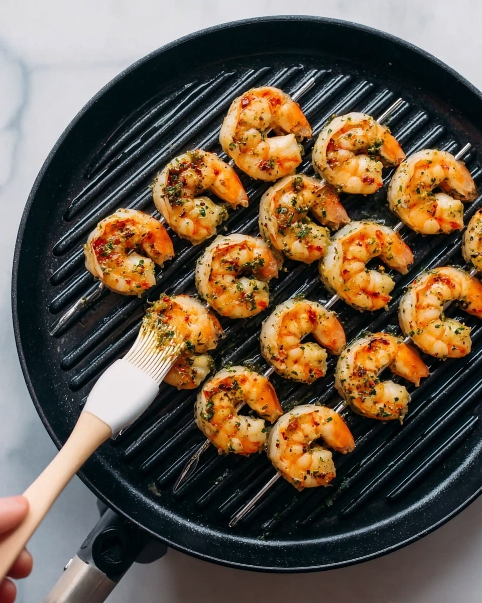 Two skewers of cooked shrimp sit on a black round grill pan with ridges. The shrimp are orange with specks of green and red herbs and spices on the surface. A woman's hand holds a white basting brush painting sauce or oil on the shrimp. The pan is on a white marbled surface. photo taken with an iphone --ar 4:5 --v 7
