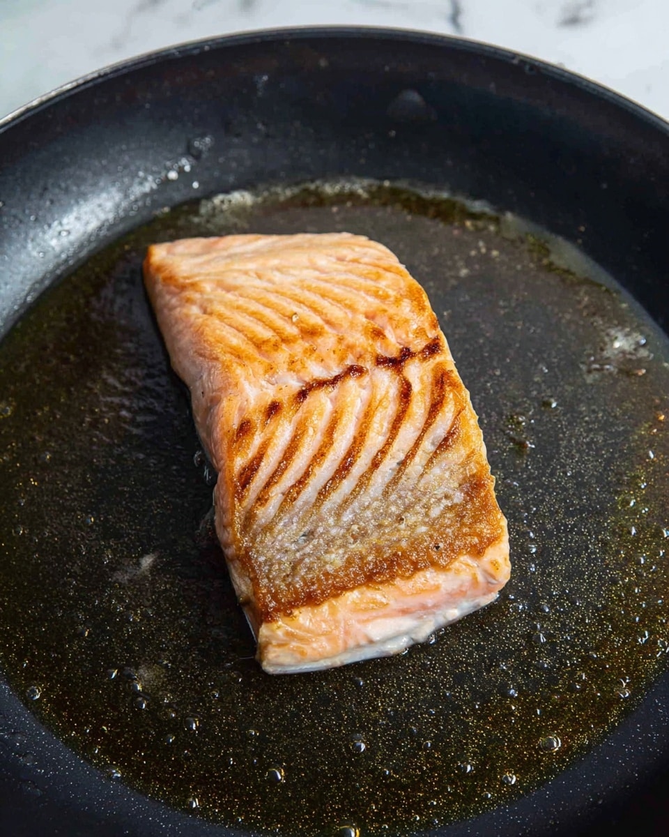 A single fillet of salmon is cooking in a large black pan with a shiny layer of oil. The salmon is positioned in the center, showing a golden brown crispy skin on top with slight darker grill marks. The visible side of the salmon has a pale pink color with a soft texture, and small droplets of oil can be seen around it on the pan's surface. The background is a white marbled texture. photo taken with an iphone --ar 4:5 --v 7