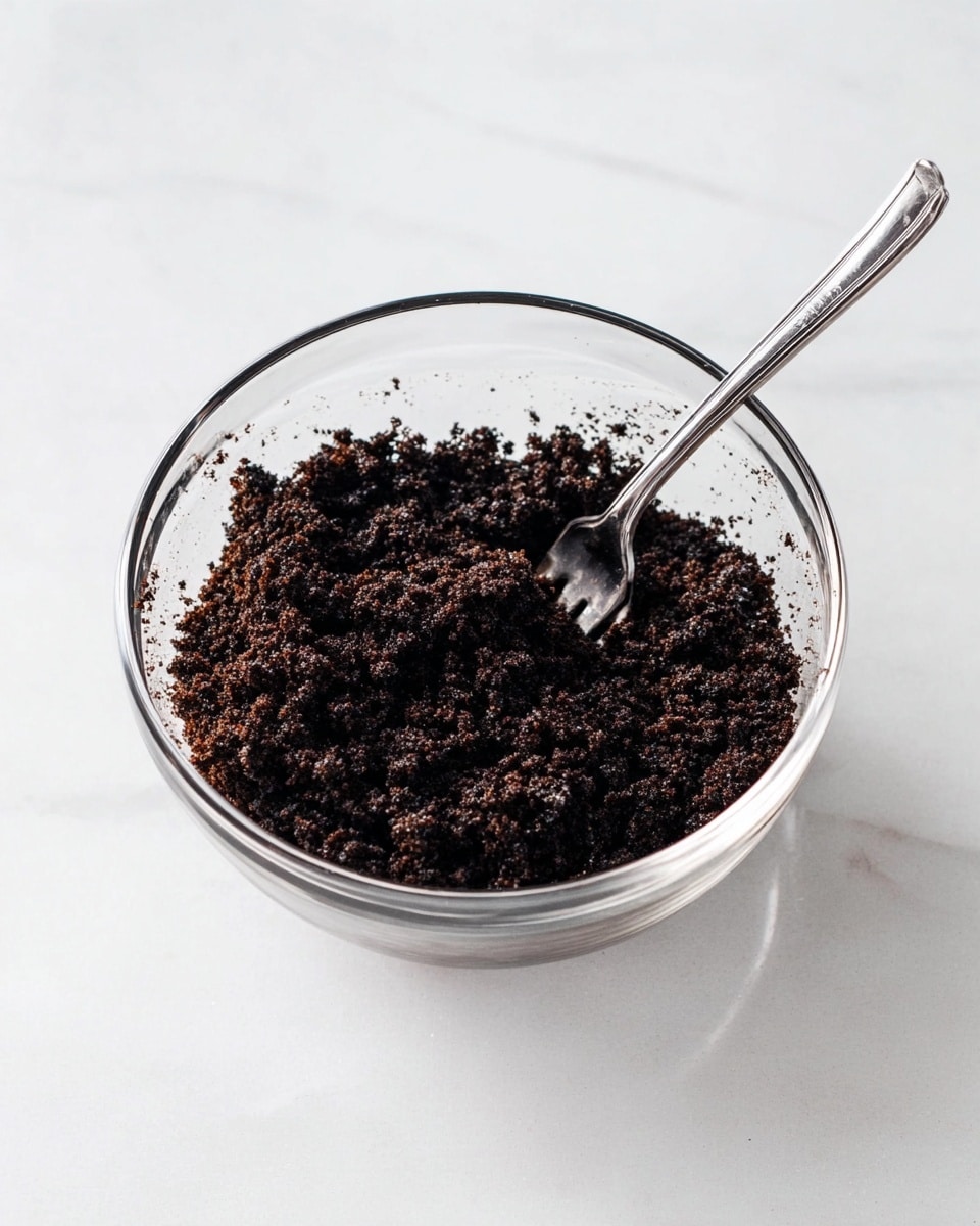 A clear glass bowl sits on a white marbled surface, filled with dark brown crumbly mixture that looks soft and grainy. A silver fork rests inside the bowl, its prongs partially covered by the dark crumbs, positioned at a slight angle toward the top edge of the bowl. The crumb mixture is evenly spread in the bowl with some small clusters and loose bits around the sides. The overall scene is clean and bright with a simple, neat look photo taken with an iphone --ar 4:5 --v 7