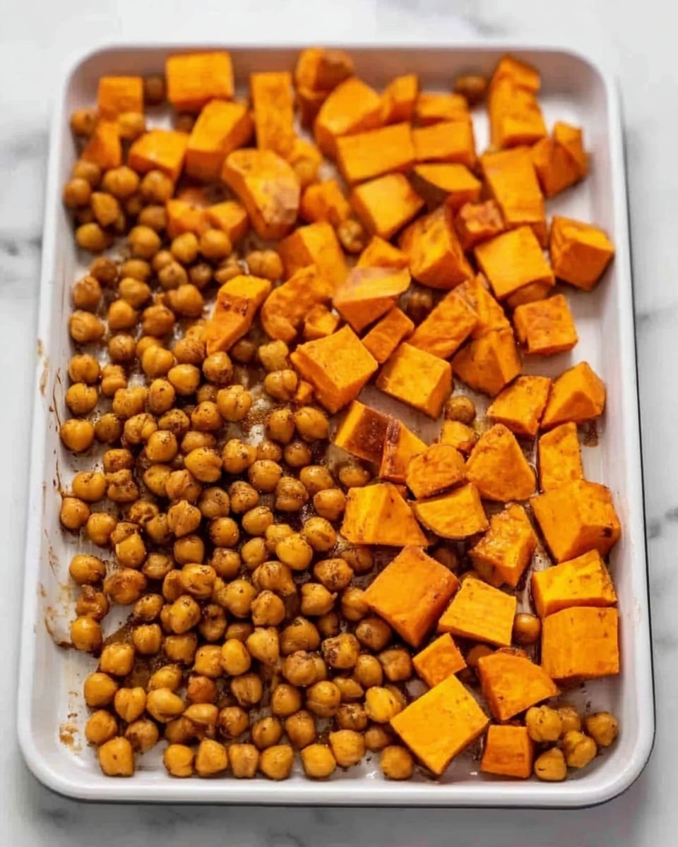 The image shows a white baking tray filled with two layers of food on a white marbled surface. The bottom layer consists of golden-brown roasted chickpeas, rounded and lightly textured. Above them, there are chunky pieces of bright orange roasted sweet potatoes, some with slightly darker roasted edges, scattered evenly across the tray. The colors contrast nicely with the white tray and marbled background, creating a warm and inviting look. photo taken with an iphone --ar 4:5 --v 7