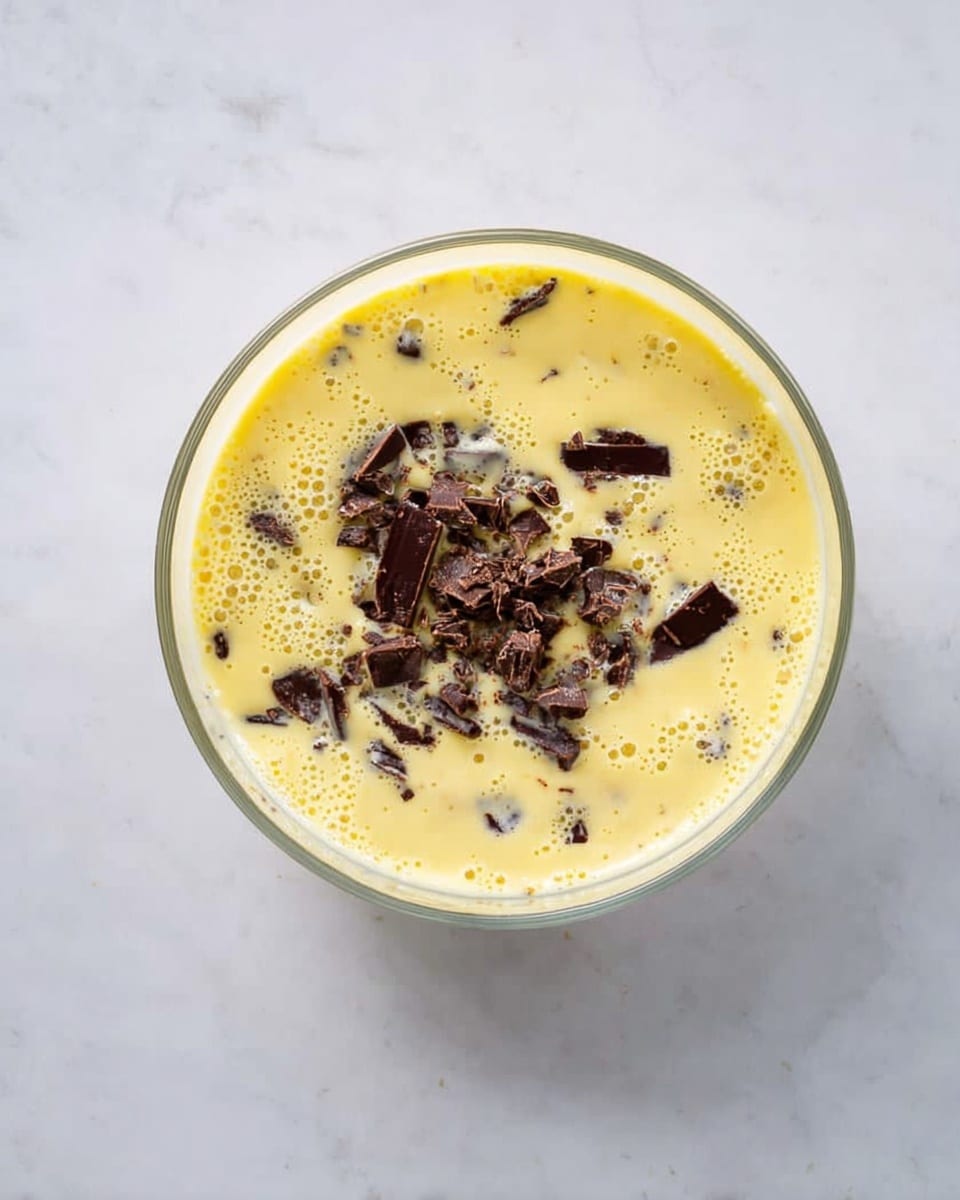 A clear glass bowl on a white marbled surface holds a light yellow creamy mixture with small bubbles, filled with uneven dark brown chocolate chunks spread on the top surface and partially submerged. The bowl is viewed from above, showing the smooth texture of the mixture with scattered pieces. photo taken with an iphone --ar 4:5 --v 7