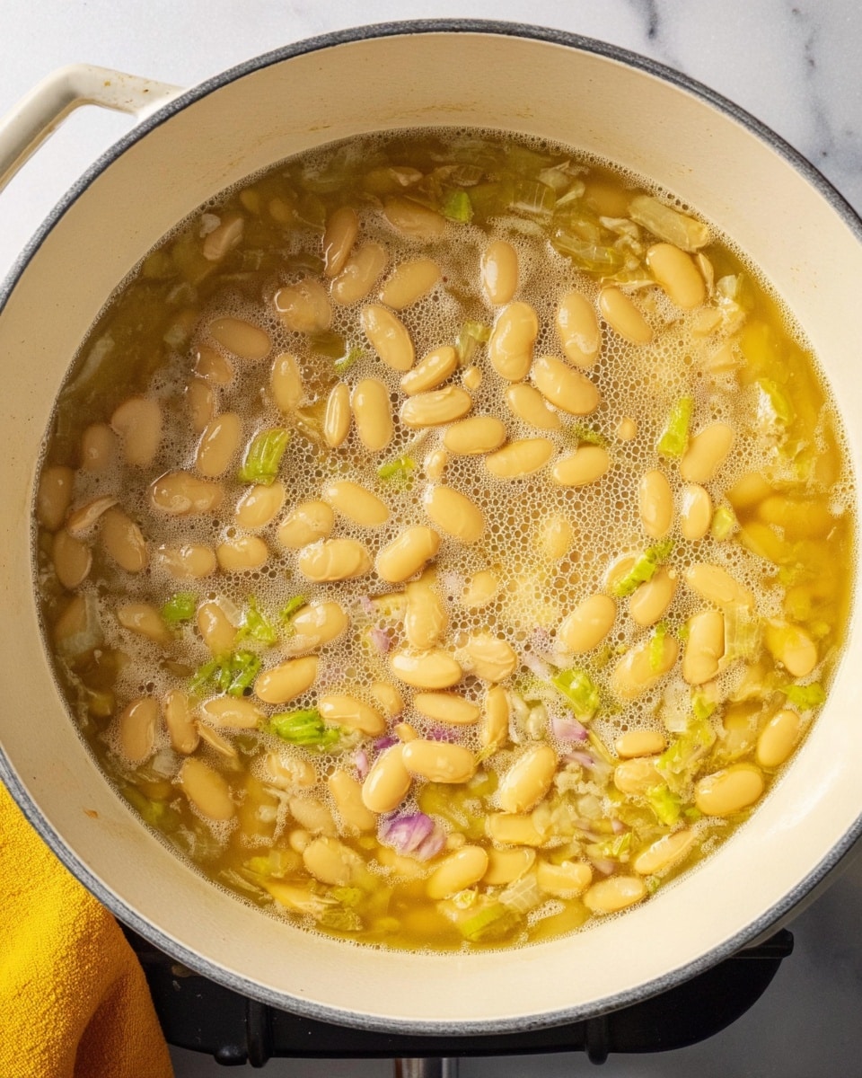 A top view of a white deep pot filled with soft yellow beans cooking in light yellow broth with small bits of green and purple vegetables mixed in. The pot is set on a stove with a white marbled surface around it and a yellow cloth at the edge. The liquid is bubbling gently over the beans, showing some foam on the surface. Photo taken with an iphone --ar 4:5 --v 7
