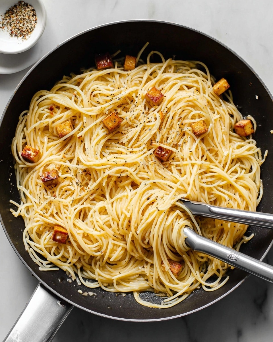 The image shows a large black pan filled with a single layer of plain spaghetti pasta that is light yellow in color. Scattered within the spaghetti are small cubes of browned tofu or cheese, adding a contrast with their golden-brown edges. The pasta looks smooth and lightly coated with oil or butter, with black pepper sprinkled evenly on top. Two silver tongs rest inside the pan, partially lifting some spaghetti, showing its long, thin, and slightly shiny texture. The pan sits on a white marbled surface with a small white dish with seasonings visible in the corner. Photo taken with an iphone --ar 4:5 --v 7