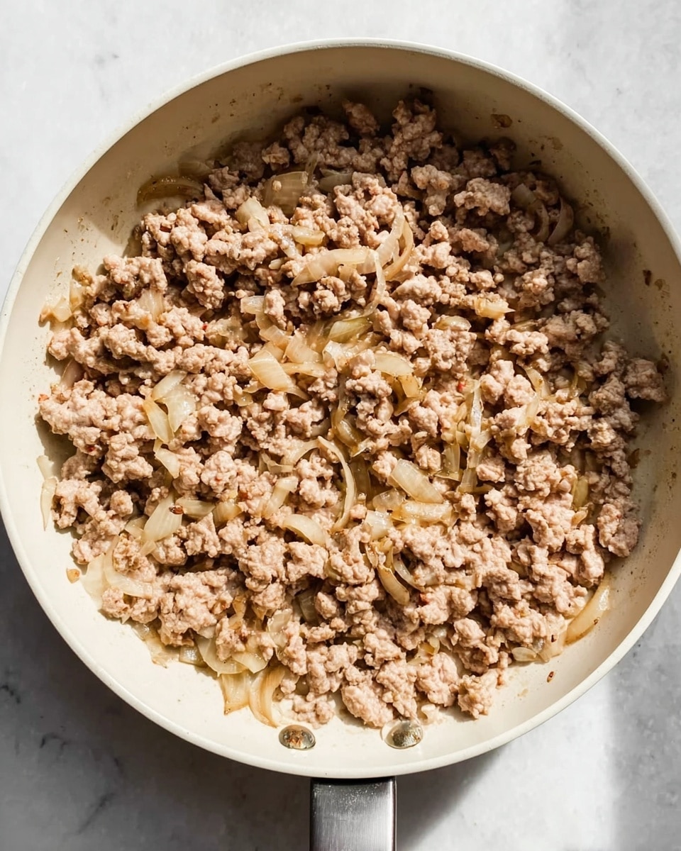 The image shows a white pan filled with cooked ground meat and small pieces of light brown, softened onions. The meat is light brown and crumbly, spread evenly across the pan with bits of translucent onions mixed throughout. The pan sits on a white marbled surface, and soft natural light shines on the dish from the side, casting gentle shadows. No other ingredients or garnishes are visible. photo taken with an iphone --ar 4:5 --v 7