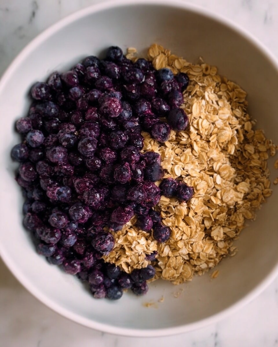 The image shows a white bowl filled with two main layers. The bottom layer is made of light golden oats with a dry and flaky texture, covering most of the bowl. On top, there is a thick layer of dark purple and blue blueberries scattered evenly, contrasting with the oat layer. The bowl sits on a surface with a white marbled texture. photo taken with an iphone --ar 4:5 --v 7