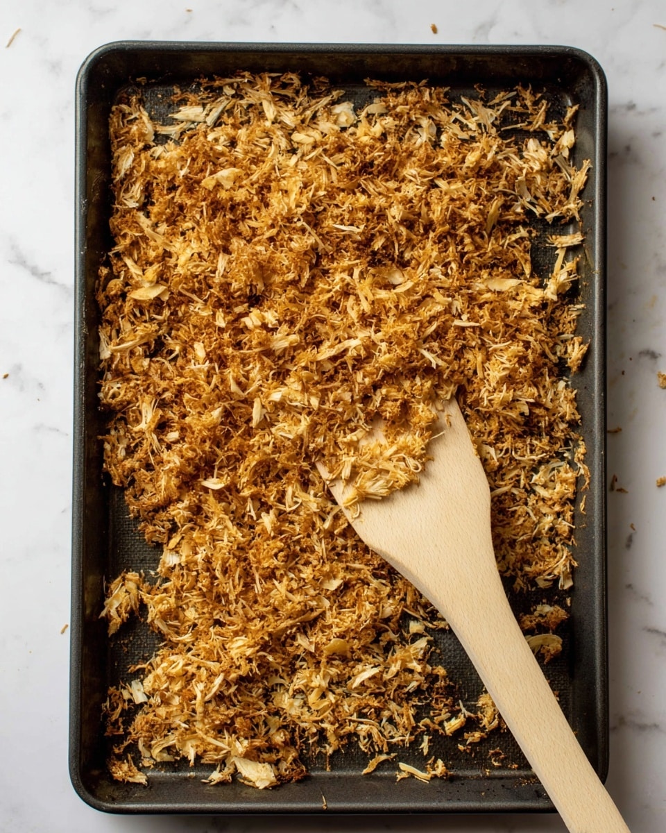 This image shows a dark baking tray filled with uneven shredded pieces of golden brown cooked food, spread across almost the entire tray surface. The shredded pieces vary in size and texture, with some parts looking crispier and darker, while others are lighter and softer. On the right side of the tray, there is a beige spatula with a wooden handle resting partially on the food, scooping up some of the shredded pieces. The tray is placed on a white marbled surface. photo taken with an iphone --ar 4:5 --v 7