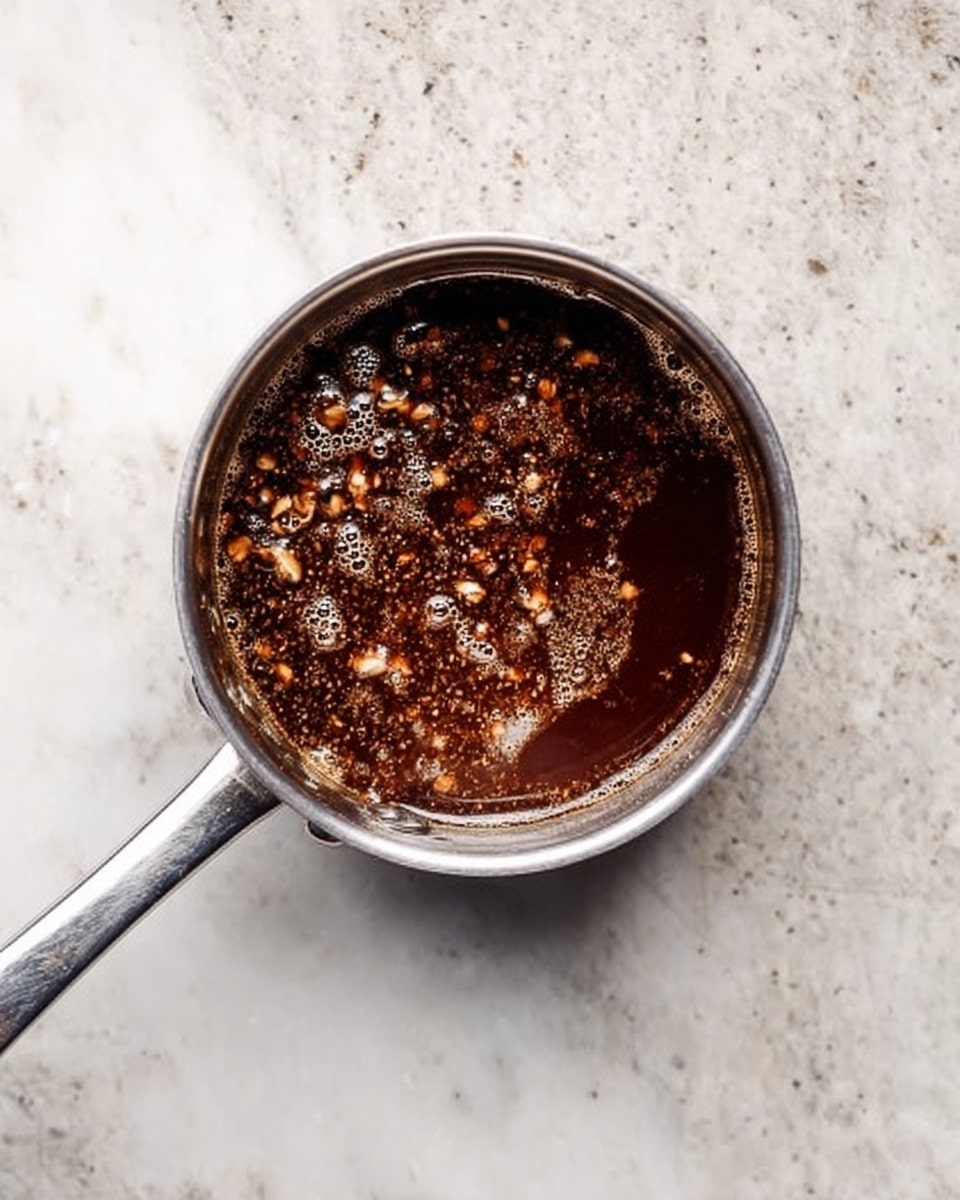 A small silver saucepan filled with dark brown liquid that has visible small pieces of nuts or spices floating inside. The saucepan rests on a white marbled surface. The liquid looks thick and glossy, with tiny bubbles spread across the top. The handle of the pan is slim and metallic, extending to the right side of the image. photo taken with an iphone --ar 4:5 --v 7