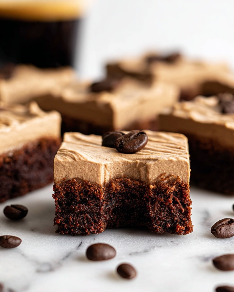 This image shows several square chocolate brownies with two layers: the bottom layer is dark and fudgy with a dense, slightly crumbly texture, and the top layer is a smooth, light brown coffee-flavored frosting with visible soft ridges on the surface. One brownie in the front has a small bite taken out of it, and two whole dark brown coffee beans are placed on top of the frosting. More coffee beans are scattered around the brownies on a white marbled surface. The background is softly blurred, and the photo has soft natural light, taken with an iphone --ar 4:5 --v 7