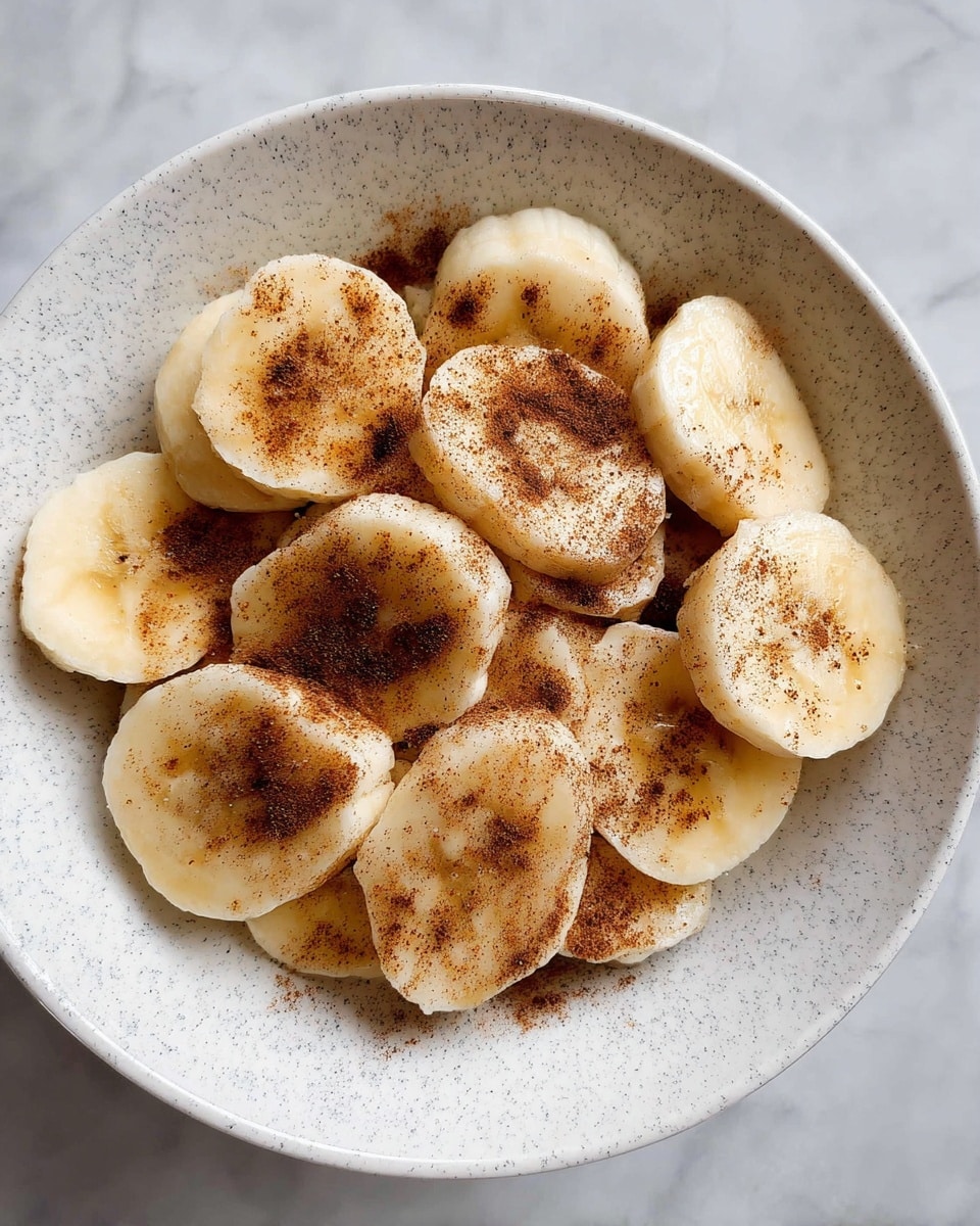 A white bowl filled with several slices of yellow banana arranged overlapping each other, topped with a light layer of dark brown cinnamon sprinkled unevenly across the banana slices, creating a contrast with the pale color of the fruit against the light grey speckled bowl and white marbled background. photo taken with an iphone --ar 4:5 --v 7