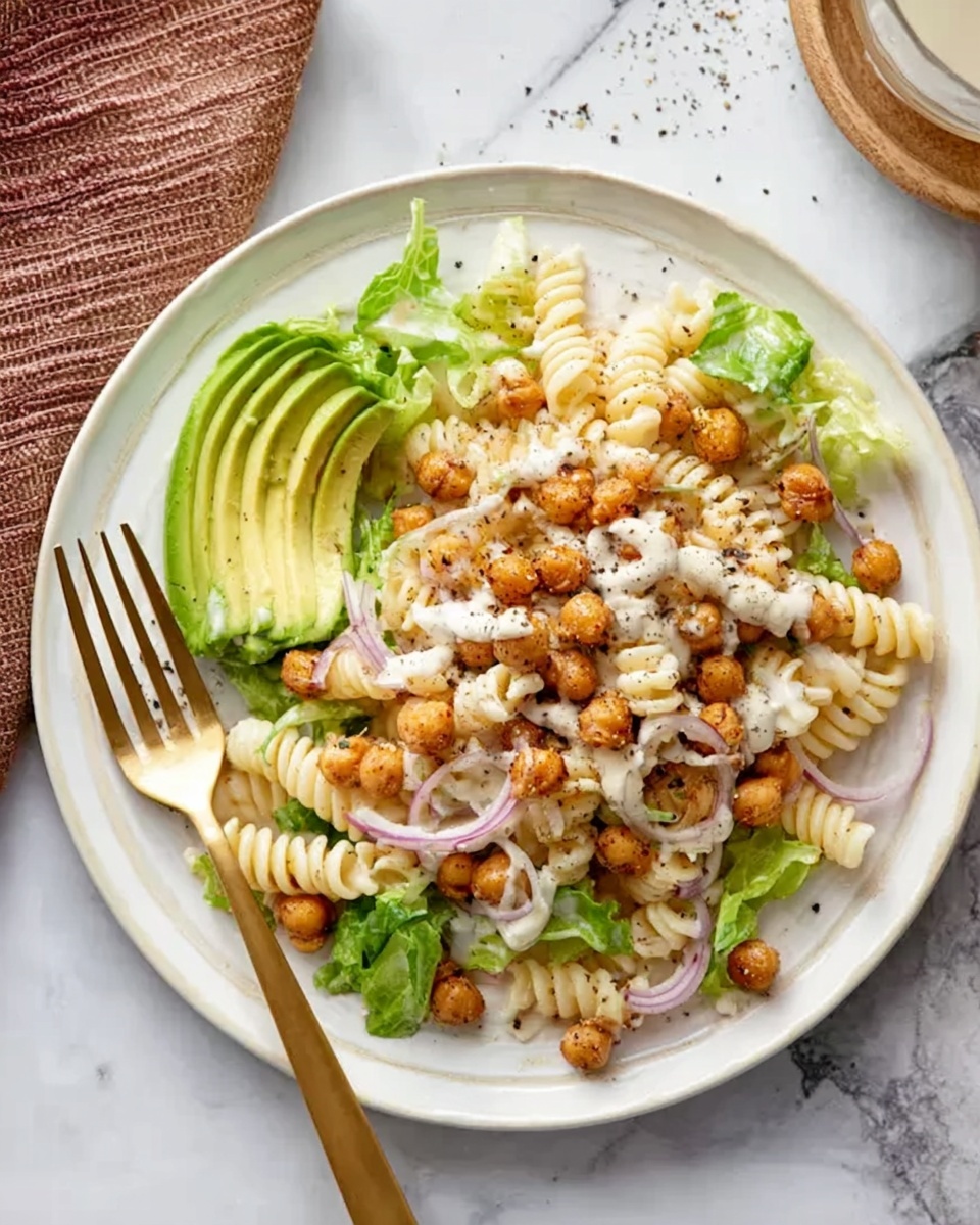 A white plate holds a pasta salad with three main layers: the base is light cream spiral pasta mixed with roasted chickpeas that are golden brown with a slightly crispy texture, scattered evenly throughout. On top of the pasta are fresh green lettuce leaves and thin slices of red onion adding pops of color. The salad is garnished with a large slice of bright green avocado resting on one side, drizzled with a creamy white dressing and sprinkled with black pepper. A gold fork rests on the left side of the plate, while the background is a white marbled surface. photo taken with an iphone --ar 4:5 --v 7