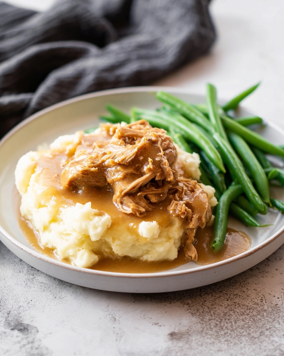 Slow Cooker Honey Mustard Chicken Recipe 5 The dish is served on a white plate placed on a white marbled surface. In the center, there is a thick layer of creamy mashed potatoes, smooth and pale yellow. On top of the mashed potatoes, there is a chunky layer of brown shredded meat covered in a light brown gravy, some gravy spilling onto the potatoes. To the right side of the plate, there is a pile of fresh, bright green steamed green beans. The overall look is warm and homey, with soft textures and natural colors. Photo taken with an iphone --ar 4:5 --v 7
