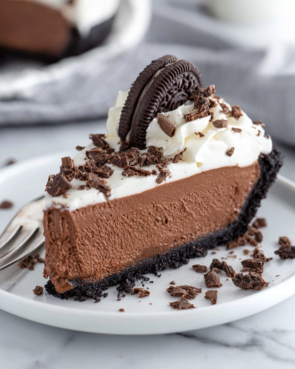 Chocolate Mousse Cake Recipe 5 A slice of chocolate mousse pie on a white plate shows three main layers: a dark, crumbly cookie crust at the bottom, a thick, smooth and airy chocolate mousse in the middle, and a topping of white whipped cream with chocolate shavings scattered on and around it. On top of the whipped cream, an Oreo cookie is placed standing up, with its dark chocolate color contrasting with the white cream filling. The plate sits on a white marbled surface and a silver fork is visible in the background near the pie. Photo taken with an iphone --ar 4:5 --v 7