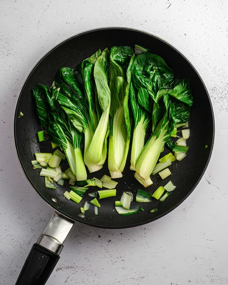 A black non-stick frying pan shows a group of roughly seven whole baby bok choy stalks in the center, their light green stems and darker green leaves spreading outward; scattered around the bok choy are small chunks of chopped white and green stalks, adding texture and color contrast. The pan handle is black with a metal section near the pan, and the pan rests on a white marbled surface. The lighting highlights the fresh, slightly wilted texture of the vegetables. photo taken with an iphone --ar 4:5 --v 7