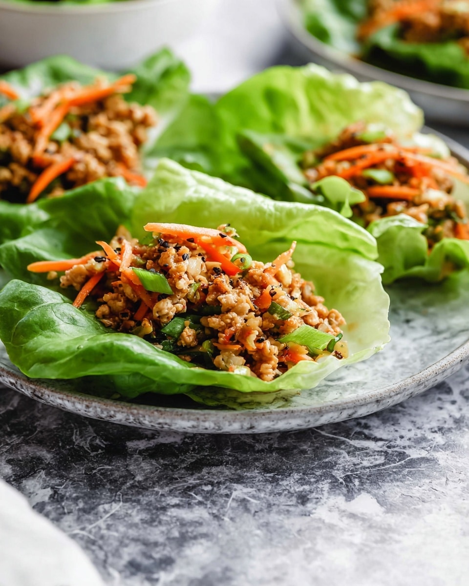A close-up view of three green butter lettuce leaves acting as cups, each filled with a mixture of small, light brown cooked pieces mixed with thin orange strips, likely carrots, and small green bits, all topped with cracked black pepper. The lettuce cups sit on a round, white plate with a gray marbled texture underneath. The background has a white marbled texture with part of another plate visible. photo taken with an iphone --ar 4:5 --v 7