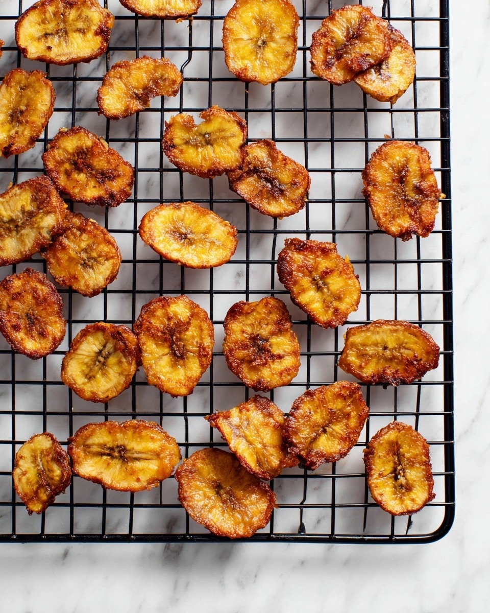 A black cooling rack holds two layers of fried plantain slices spread out with small gaps between each slice. The plantain slices are golden brown with some darker spots where they are crispier, showing a rough texture and slightly curled edges. The cooling rack is set on a white marbled surface, providing a clean and bright background that contrasts with the warm tones of the plantains. photo taken with an iphone --ar 4:5 --v 7