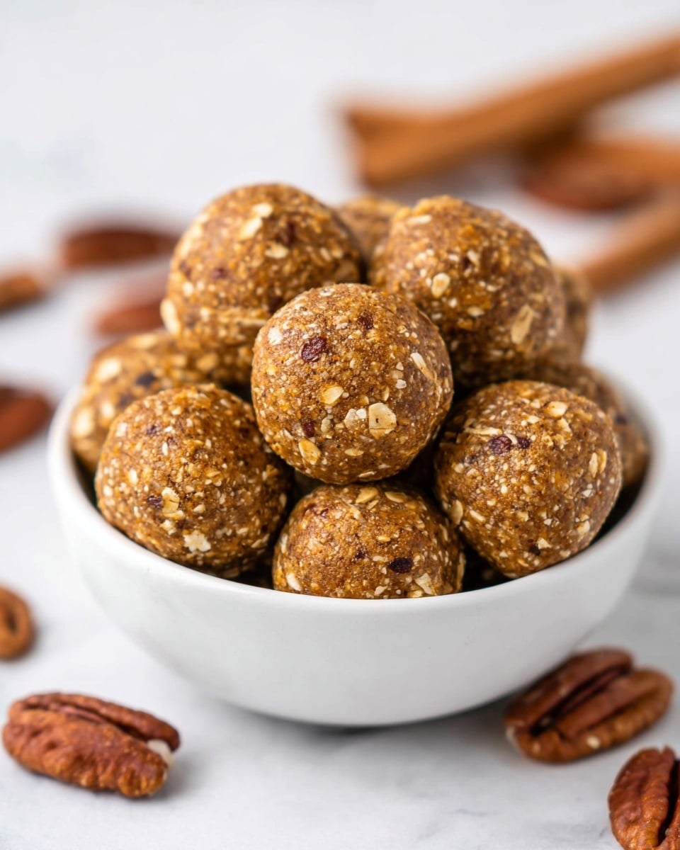 A white bowl filled with small round energy balls that have a textured surface with visible oats and small nut pieces, showing a mix of light brown and golden colors. The balls are stacked and surrounded by whole pecans, dark brown in color with a glossy texture. In the background, on a white marbled surface, there are two cinnamon sticks and blurred pecans. The focus is on the front energy ball, showing its detailed texture. Photo taken with an iphone --ar 4:5 --v 7