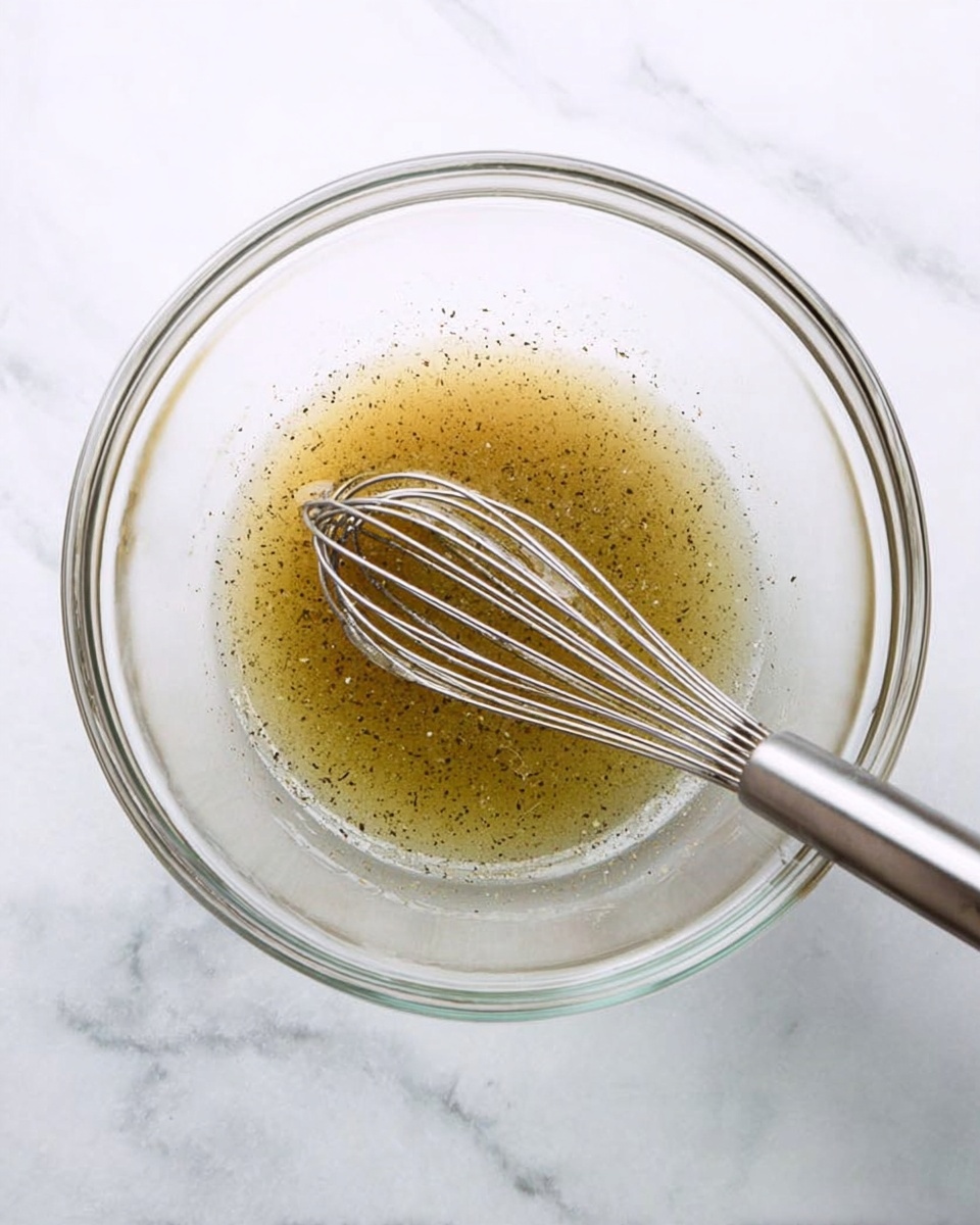 A clear glass bowl on a white marbled surface holds a thin, light brown liquid with small black specks, likely seasoning. A silver metal whisk is inside the bowl, with a woman's hand holding it from the right side of the image, stirring the mixture gently. The scene is clean and bright with soft natural light, emphasizing the smooth texture of the liquid and the glass bowl's transparency. photo taken with an iphone --ar 4:5 --v 7