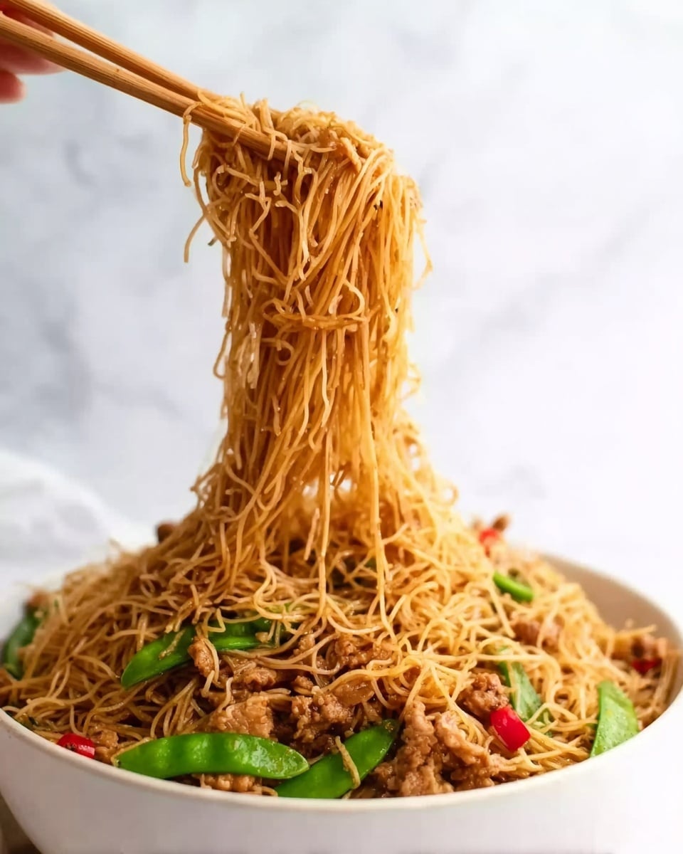 A white bowl filled with thin noodles mixed with pieces of light brown meat, green snap peas, and small red chili slices. A pair of wooden chopsticks held by a woman's hand lifts a large portion of the noodles above the bowl, showing the long, slightly shiny noodles dripping down. The background is a soft white marbled texture, giving a clean and bright look. Photo taken with an iphone --ar 4:5 --v 7