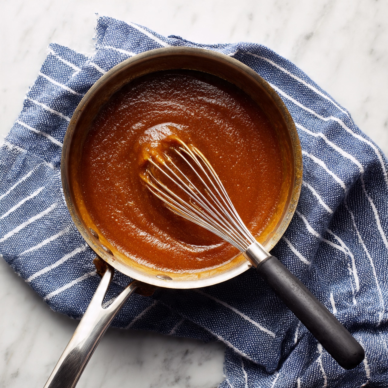 A silver metal pan with a long handle holds a thick, lumpy brown sauce with a shiny texture. A metal whisk with a black handle is resting inside the pan, partially lifted, showing the sauce clinging to the wires. The pan is placed on a white marbled surface, next to a blue and white striped cloth draped casually to the right. The light is soft, highlighting the sauce’s rough texture and glossy areas. photo taken with an iphone --ar 4:5 --v 7