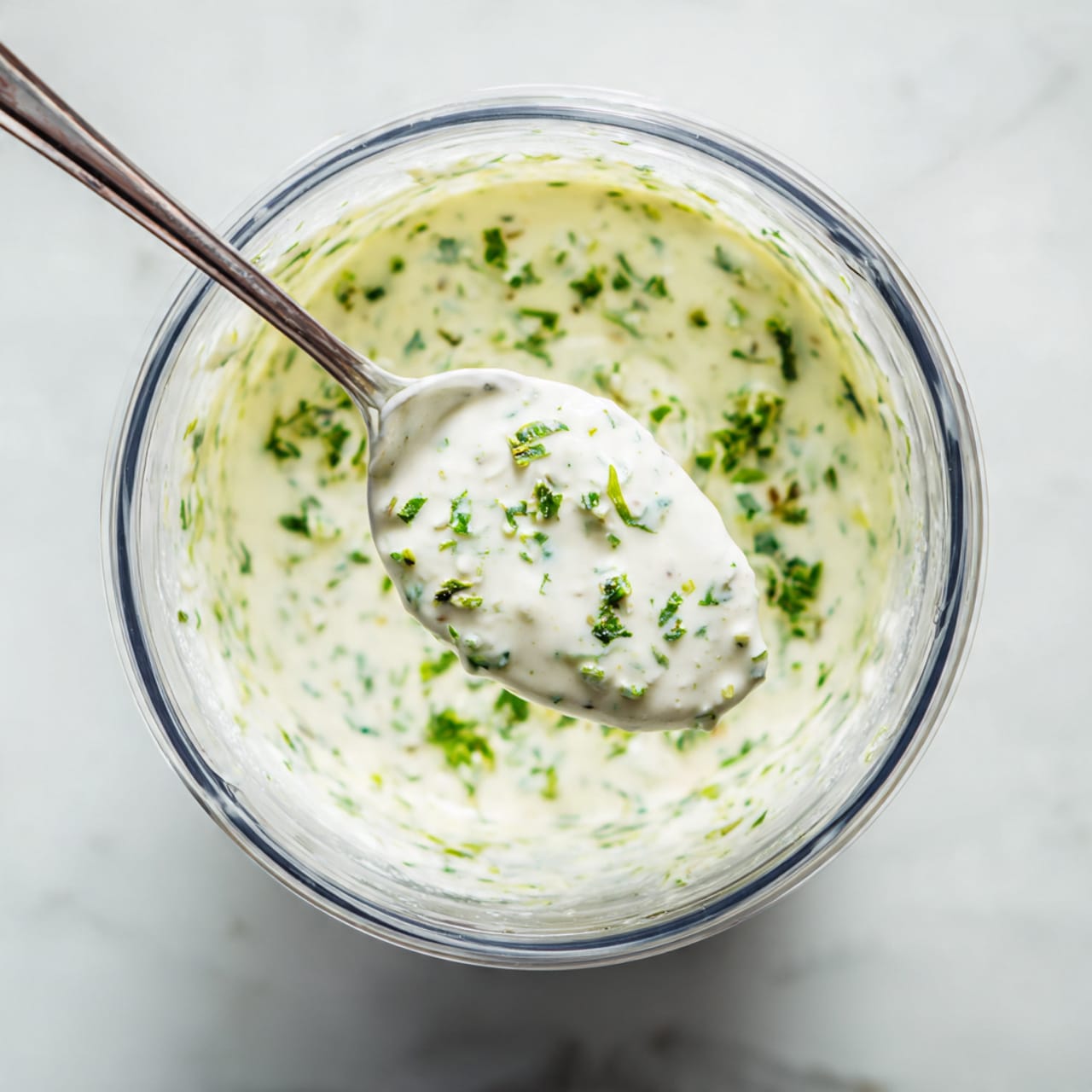 The image shows a clear blender container filled with a thick, creamy sauce that has small green herbs mixed throughout. Inside the blender, a metal spoon is lifted, holding a scoop of the thick, white sauce speckled with green bits. The blender sits on a white marbled surface. photo taken with an iphone --ar 4:5 --v 7