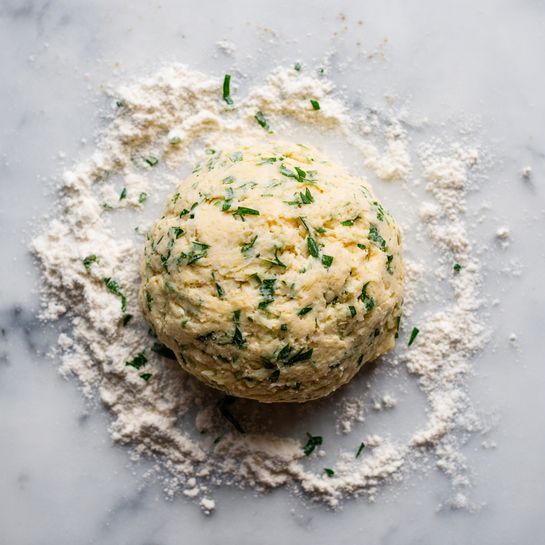 A ball of dough mixed with green herbs is placed in the center of a white marbled surface. The dough is pale beige with small green specks spread all over, giving it a slightly textured look. Light dustings of white flour surround the dough, blending softly on the marble. The dough has a soft, slightly uneven round shape, showing it is fresh and ready to be used. photo taken with an iphone --ar 4:5 --v 7