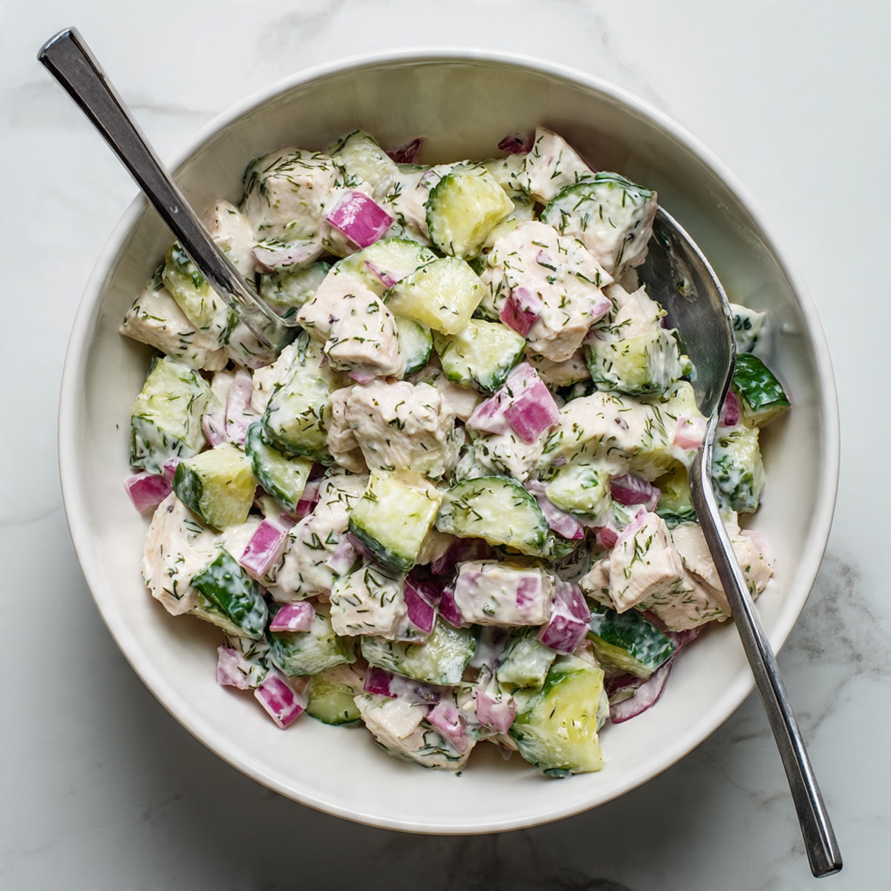 A large white bowl filled with a mixed salad that has three main layers: light pink chunks of cooked chicken, small green cucumber pieces, and tiny bits of red onion, all coated with a creamy white dressing mixed with flecks of green herbs. Two gray-handled spoons rest inside the bowl, one on the left and one on the right, slightly covered with the salad. The bowl sits on a white marbled surface with a clean and bright background. photo taken with an iphone --ar 4:5 --v 7