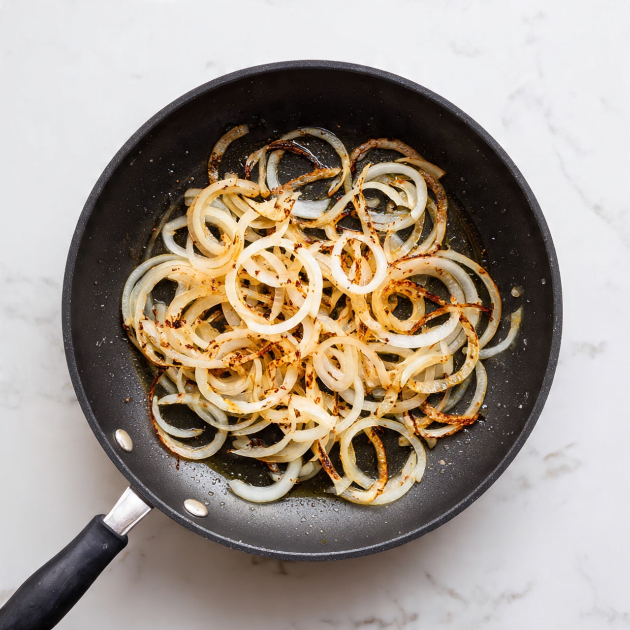A black frying pan holds a single layer of thinly sliced sautéed onions. The onions are light golden with some edges browned and crispy, showing a mix of round rings and long strips spread unevenly across the pan. Small puddles of oil glisten around the onions. The frying pan is set on a stove with a white marbled surface visible at the bottom right corner. photo taken with an iphone --ar 4:5 --v 7
