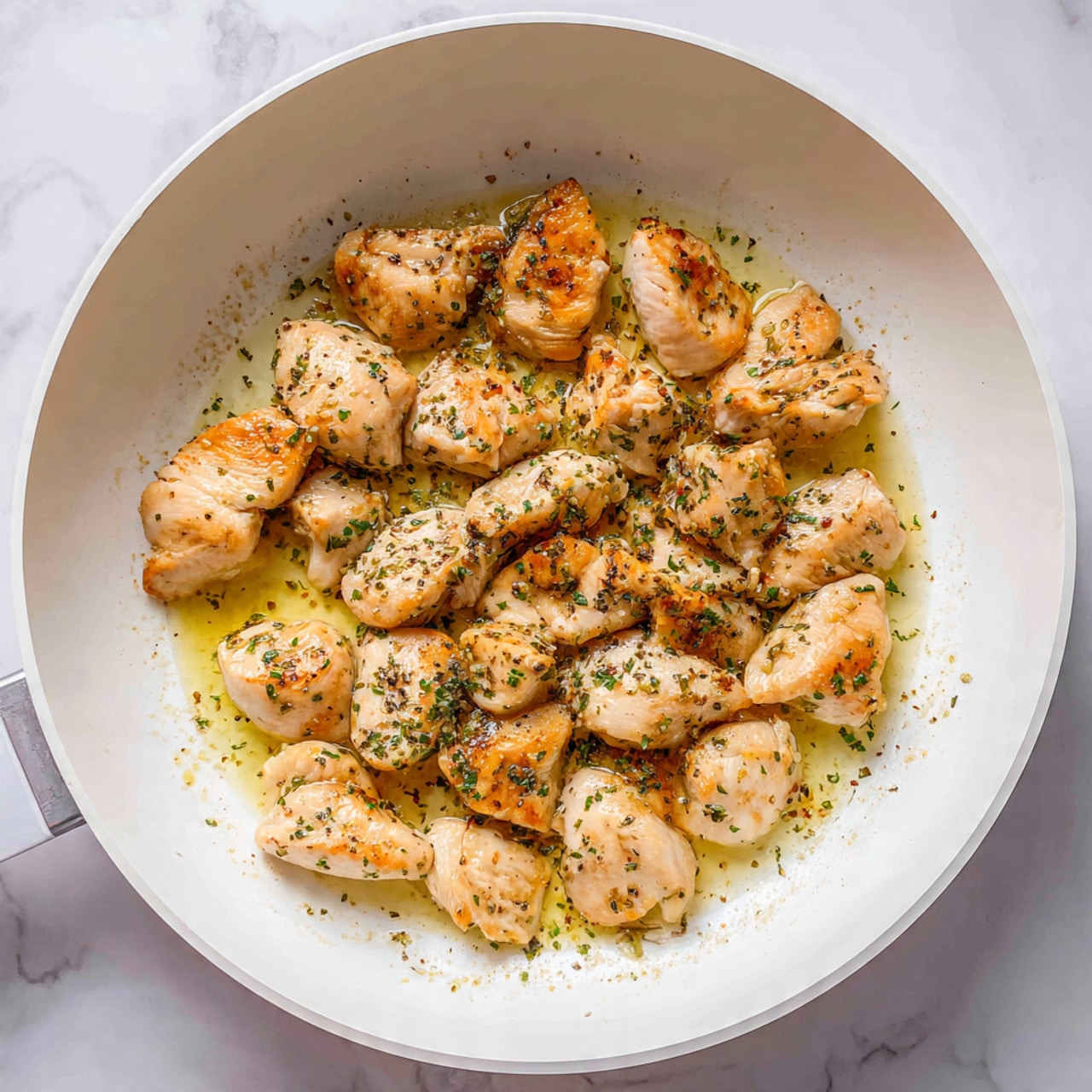 The image shows a white pan with several small pieces of chicken cooking inside. The chicken pieces are light golden brown on some parts, with a slightly soft texture on others, showing a mix of cooked and partly raw areas. Small specks of black pepper and green herbs are sprinkled evenly over the chicken, with a thin layer of oil making the surface slightly shiny. The pan is placed on a white marbled surface. photo taken with an iphone --ar 4:5 --v 7