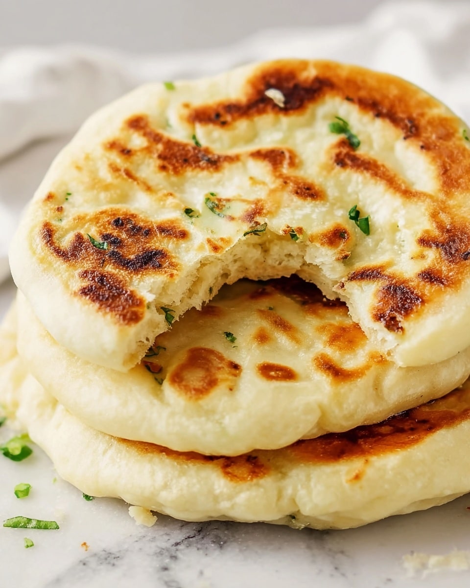 A stack of three round flatbreads with a soft, fluffy texture sits on a white marbled surface. The flatbreads are light golden in color with darker brown, uneven toasted spots scattered across the top layers. The top flatbread is partially torn, showing a thick, pillowy inside. Small pieces of green herbs peek out slightly between the flatbreads. The image has a bright and clean look. photo taken with an iphone --ar 4:5 --v 7