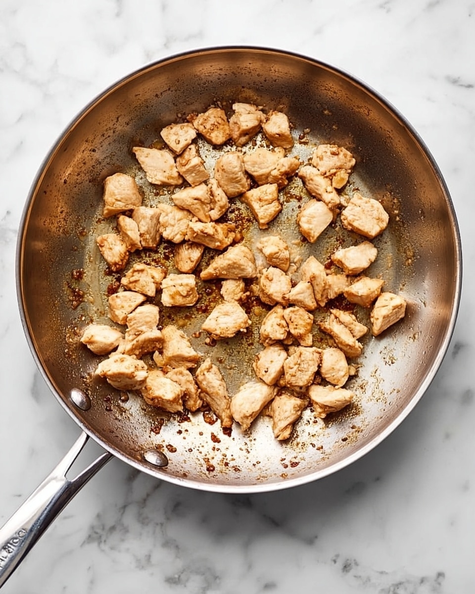 A stainless steel frying pan is shown from above on a white marbled surface, filled with small, unevenly shaped pieces of light brown cooked chicken scattered across the bottom. The chicken pieces have a slightly crispy texture with some browned spots. The pan's interior shows some browned bits and light oil residue, giving it a well-used look, and its metal handle extends out to the left side. Photo taken with an iphone --ar 4:5 --v 7