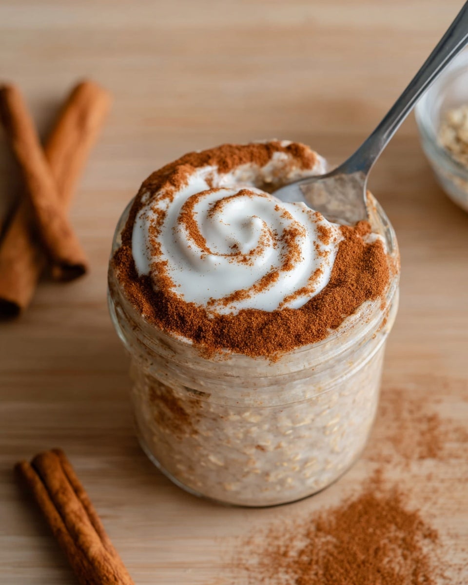 A small clear glass jar filled with oatmeal that has a light beige color and soft texture, topped with a thick layer of brown cinnamon powder evenly spread on top. Over the cinnamon, there is a white creamy swirl drizzle in a spiral pattern. A silver spoon is partly inside the jar scooping some oatmeal. The jar is on a warm light wooden surface with two cinnamon sticks nearby. Photo taken with an iphone --ar 4:5 --v 7