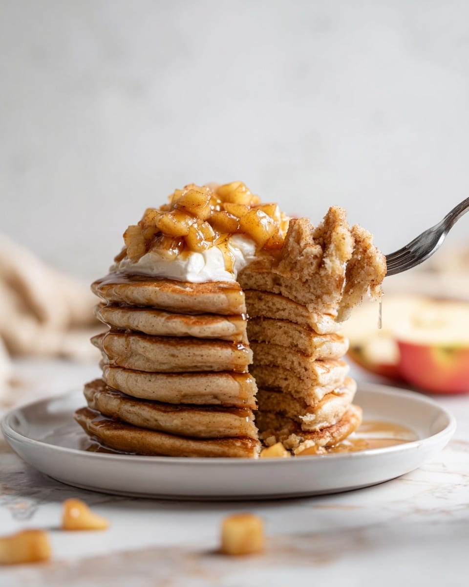 A tall stack of seven light brown pancakes sits centrally on a white plate, topped with a dollop of white cream and a generous layer of small, golden brown apple pieces mixed with cinnamon. The pancakes have a soft, slightly porous texture, and golden syrup glistens down the sides, pooling slightly on the plate. A fork is holding a bite-sized piece of pancake from the middle layers on the right side. There are a few apple pieces scattered on the white marbled surface below the plate. The background is softly blurred with light tones, highlighting the stack in the foreground. photo taken with an iphone --ar 4:5 --v 7