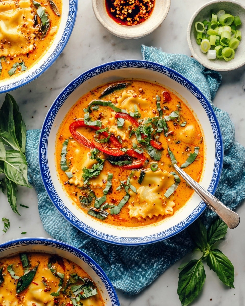 The image shows two white bowls with a blue rim, each filled with an orange, creamy soup with visible layers of ravioli pasta with wavy edges, thin strips of red bell pepper, and green herbs like chopped cilantro and basil scattered on top. A silver spoon rests inside the front bowl, which sits partially on a blue and white cloth. Surrounding the bowls on a white marbled surface are small white bowls filled with sliced green onions and a dark chili oil with chili flakes. Fresh basil leaves and scattered herb pieces add fresh green color around the soup bowls. Photo taken with an iphone --ar 4:5 --v 7