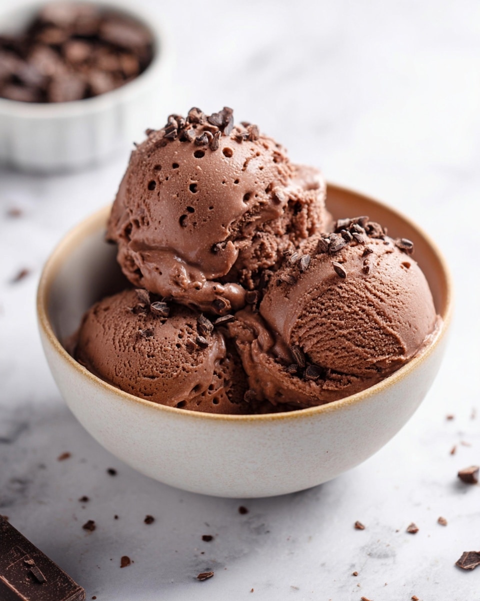The image shows three scoops of rich chocolate ice cream placed closely together in a white bowl. The ice cream scoops have a creamy, smooth texture with tiny holes and soft melting edges. Sprinkled on top and around the ice cream are small dark chocolate pieces adding texture and contrast. The bowl sits on a white marbled surface, with some scattered chocolate bits nearby. In the background, there is a blurred white bowl filled with more chocolate chunks. Photo taken with an iphone --ar 4:5 --v 7