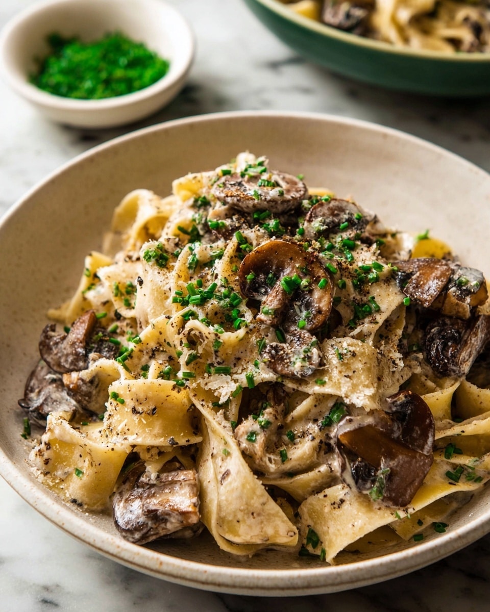 A close-up of a bowl of pasta with creamy mushroom sauce, showing wide noodles mixed thoroughly with sliced brown mushrooms coated in a light cream sauce. The pasta has a soft beige color with specks of black pepper sprinkled all over. The dish is topped with finely chopped green chives adding a fresh pop of color. The noodles and mushrooms are piled in a shallow white bowl, set on a white marbled surface, with a small white bowl of green herbs blurred in the background. Photo taken with an iphone --ar 4:5 --v 7