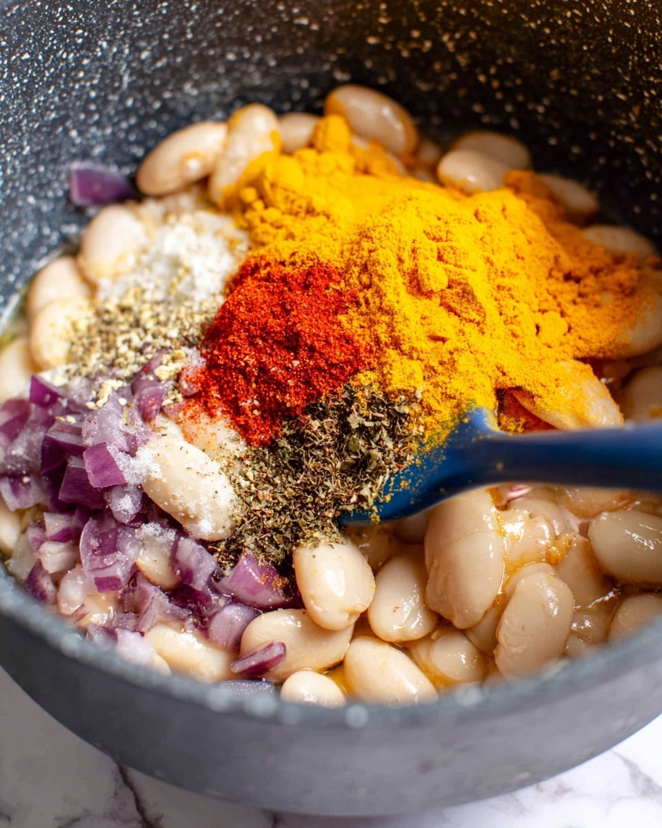 Inside a dark grey pot with speckled texture, there are several layers of ingredients: a base layer of cooked white beans, creamy white liquid around the beans, and small pieces of cooked purple onion scattered along the edges. On top of the beans, there is a mound of vibrant yellow turmeric powder, a heap of bright red paprika, white granulated salt, and various black and green mixed dried herbs and spices. A blue silicone spatula is partially visible, lifting some of the spices and beans. The background is a white marbled texture photo taken with an iphone --ar 4:5 --v 7