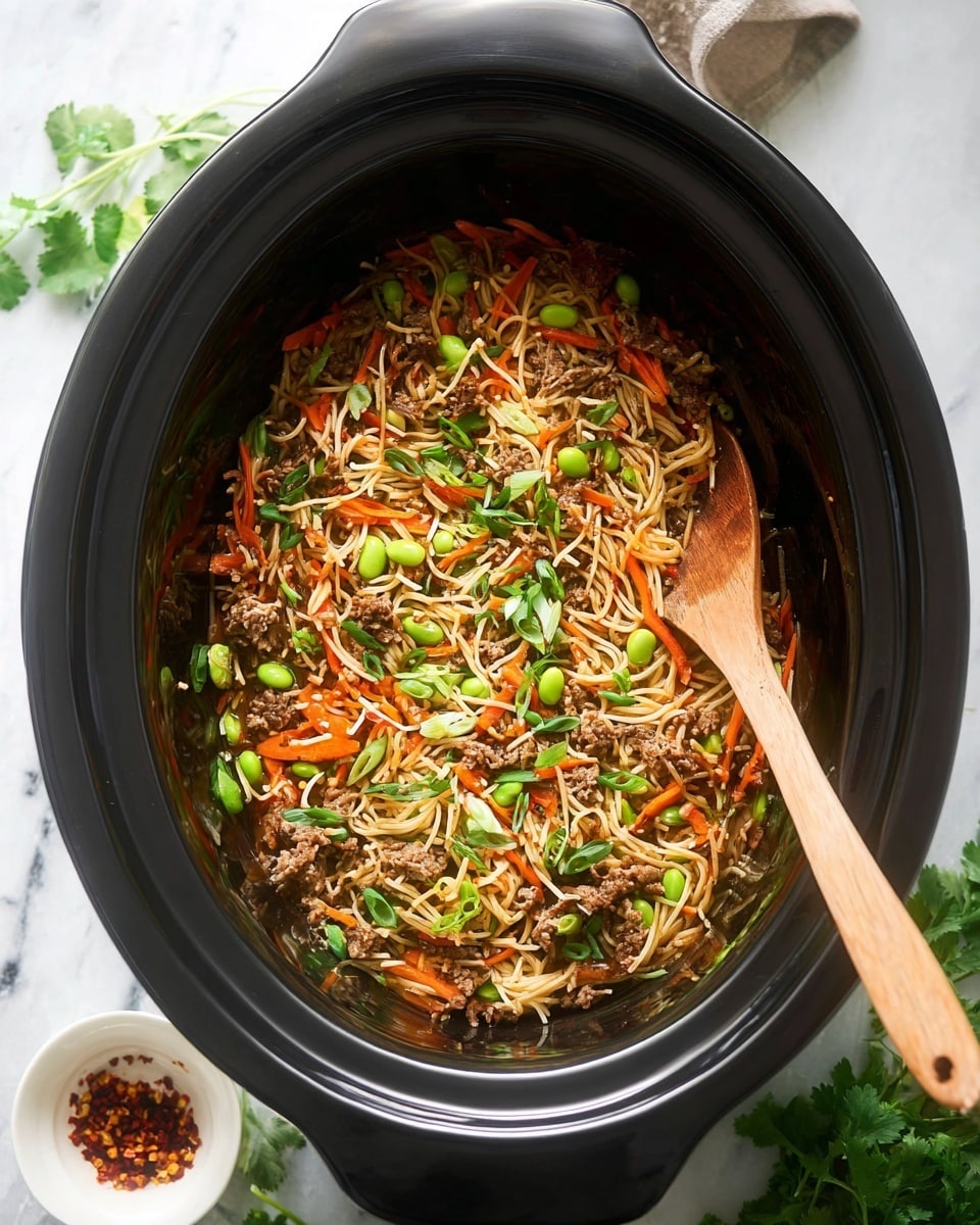 A black slow cooker filled with thin noodles mixed with small pieces of cooked meat, thin orange carrot strips, green edamame beans, and chopped green onions scattered on top. There are also bits of fresh green cilantro leaves spread around. A light wooden spoon is placed inside the cooker, resting over the noodles on the right side. The cooker sits on a white marbled surface with some green herb leaves and a small white bowl with red chili flakes visible in the background photo taken with an iphone --ar 4:5 --v 7