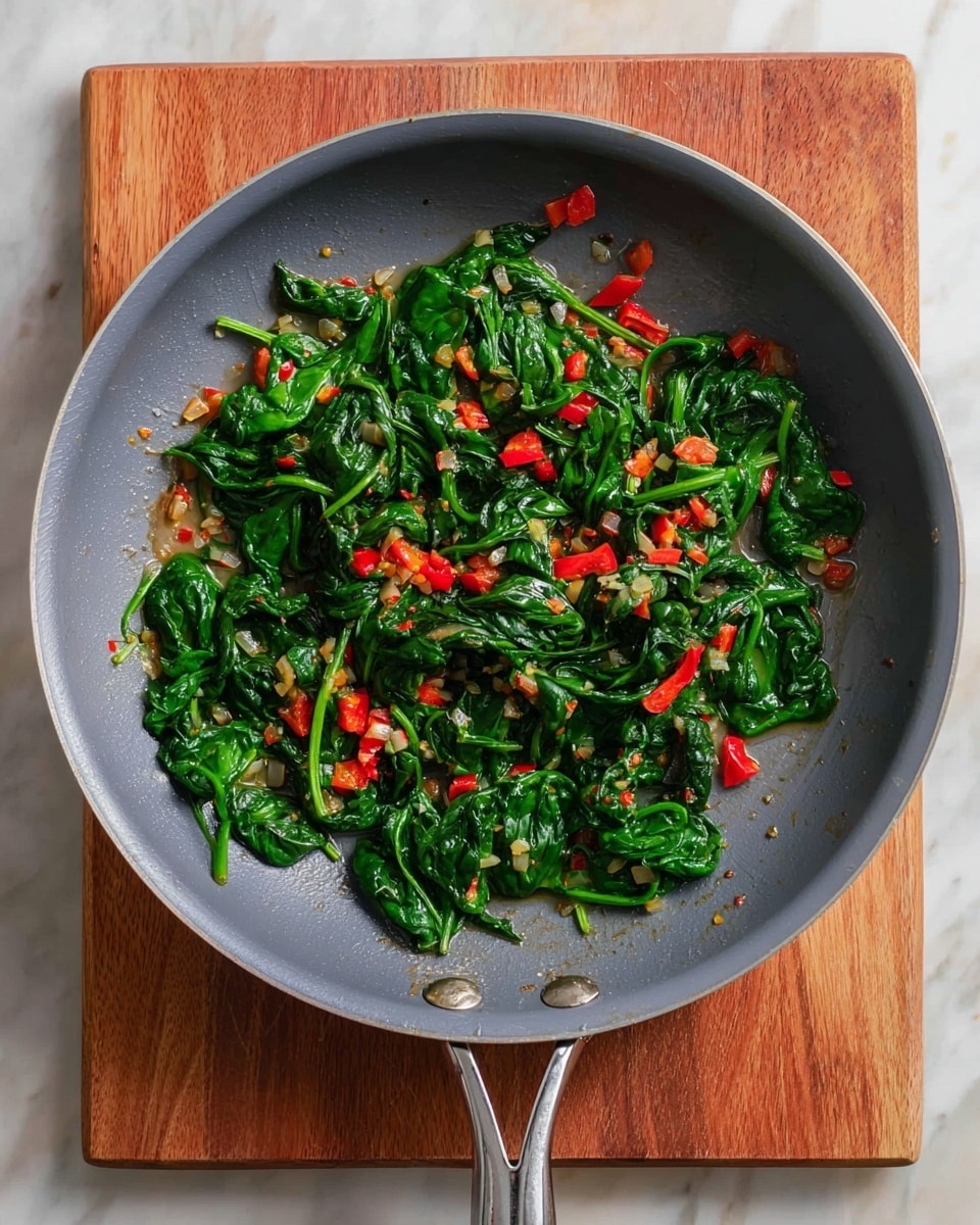 A gray frying pan filled with cooked spinach leaves and small pieces of red bell pepper mixed with tiny bits of cooked onion. The spinach leaves are bright green and soft, spread evenly across the pan. The red bell pepper adds bright red color, scattered on top and mixed throughout. The onion pieces are pale yellow and small, providing texture and color contrast. The pan has a shiny metal handle and sits on a wooden cutting board. The background is a white marbled surface. photo taken with an iphone --ar 4:5 --v 7