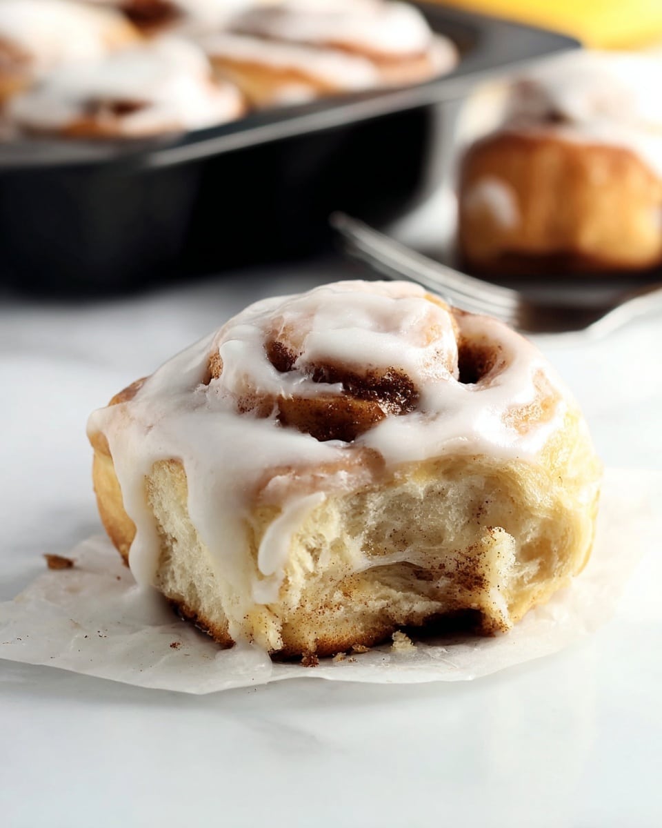 A close-up view of a cinnamon roll with one visible layer of soft, golden-brown dough swirled with a darker cinnamon filling. The top is thickly covered with white creamy icing, dripping slightly over the edges, giving it a smooth, shiny texture. The cinnamon roll sits on a piece of white parchment paper on a white marbled surface. In the soft-focused background, some more cinnamon rolls can be seen inside a black baking pan and one more cinnamon roll to the right. Photo taken with an iphone --ar 4:5 --v 7