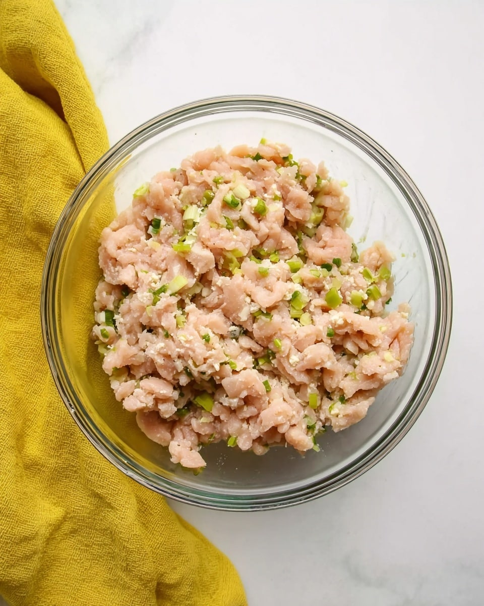 A clear glass bowl filled with light pink raw ground chicken mixed with small bits of light green chopped scallions sits on a white marbled surface. A soft yellow cloth is placed to the left side of the bowl, adding color to the scene. The texture of the meat looks soft and slightly wet, with small garlic pieces mixed in evenly. The image is simple and clean, showing the bowl from above. photo taken with an iphone --ar 4:5 --v 7