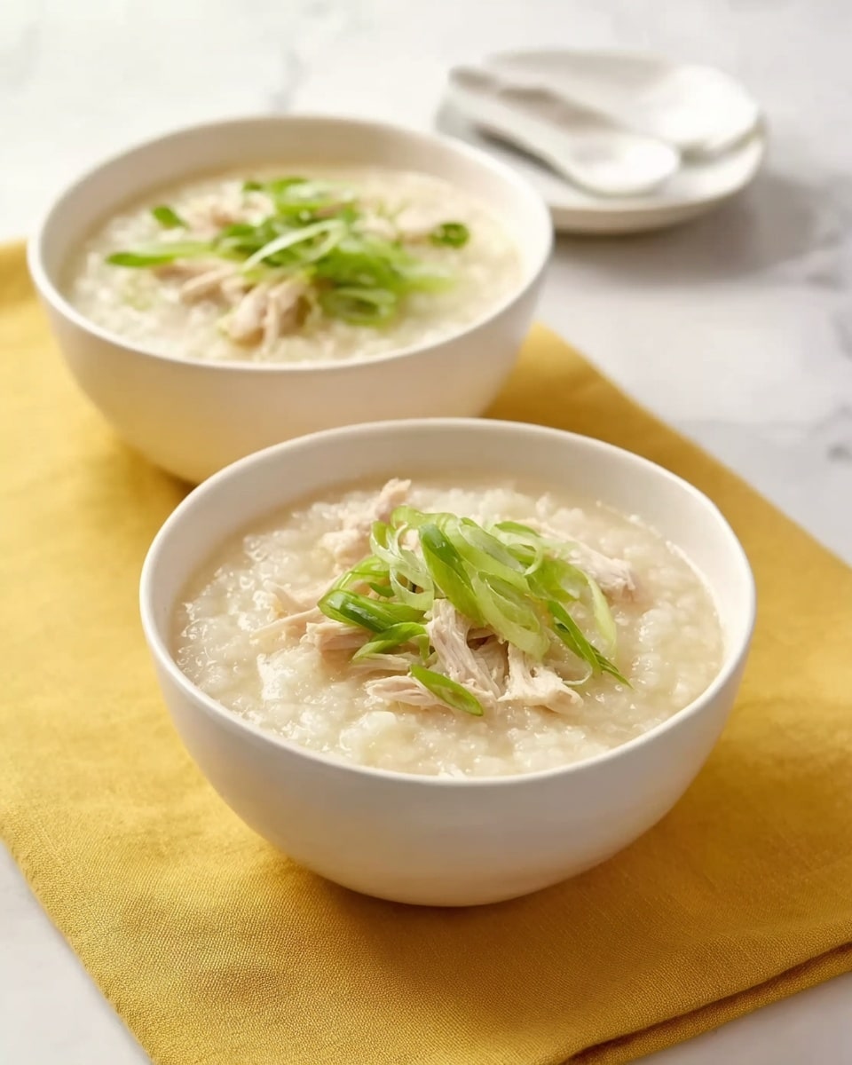 The image shows two white bowls filled with creamy rice porridge, topped with thin green slices of scallion and light shredded chicken on the surface of each bowl. Both bowls sit on a smooth yellow cloth, and there is a white ceramic spoon and a small white plate placed on a white marbled surface in the background. The porridge looks soft and thick, with a light beige color. Photo taken with an iphone --ar 4:5 --v 7