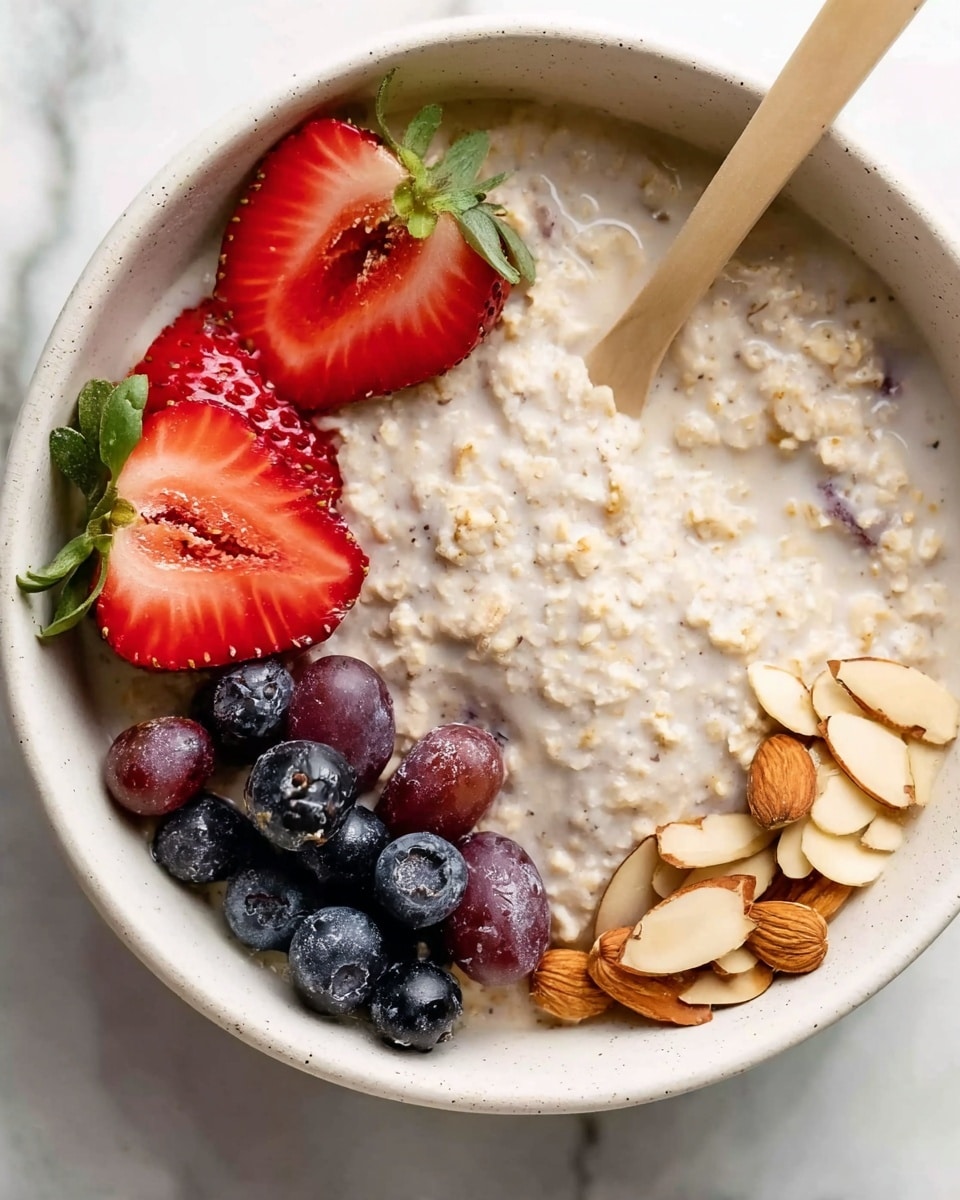 The image shows a white bowl filled with creamy oatmeal as the base layer, which has a soft texture with small grains visible. On top of the oatmeal to the upper left, there are two bright red strawberry halves with green leaves still attached. To the right of the strawberries, there is a small pile of blueberries and dark grapes with a smooth, glossy look. At the bottom right corner, thin slices of light brown almonds are scattered, showing their smooth and slightly rough edges. A wooden spoon with a light beige handle is dipped into the oatmeal on the left side of the bowl. The bowl is placed on a white marbled surface. photo taken with an iphone --ar 4:5 --v 7