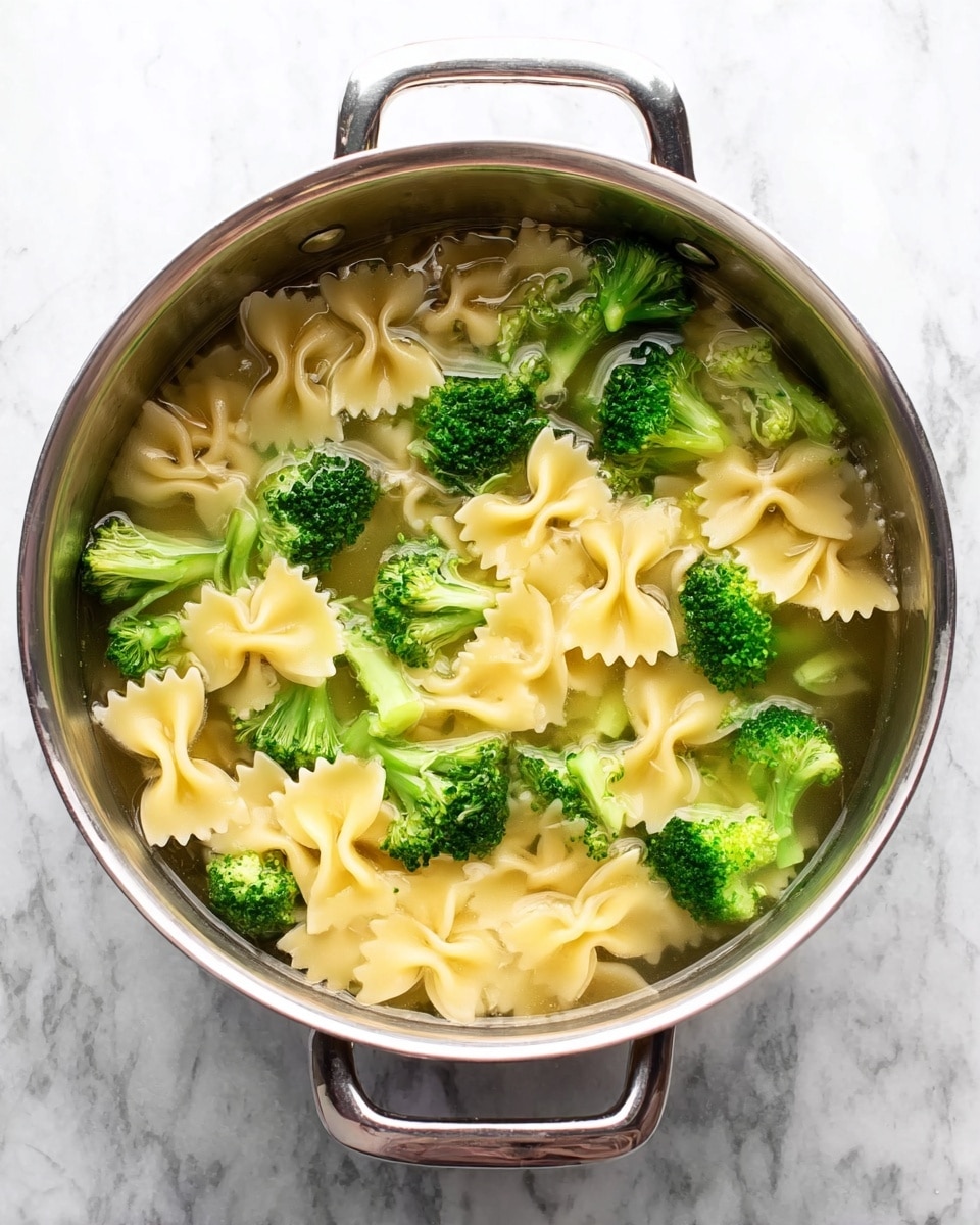 A shiny metal pot filled with water soaking light yellow bow-tie pasta and bright green broccoli florets, all floating together with some pasta pieces slightly overlapping the broccoli. The pot sits on a white marbled surface, showing its round shape and two handles on each side. The water is clear, allowing a full view of the smooth pasta and textured broccoli, both evenly mixed. Photo taken with an iphone --ar 4:5 --v 7