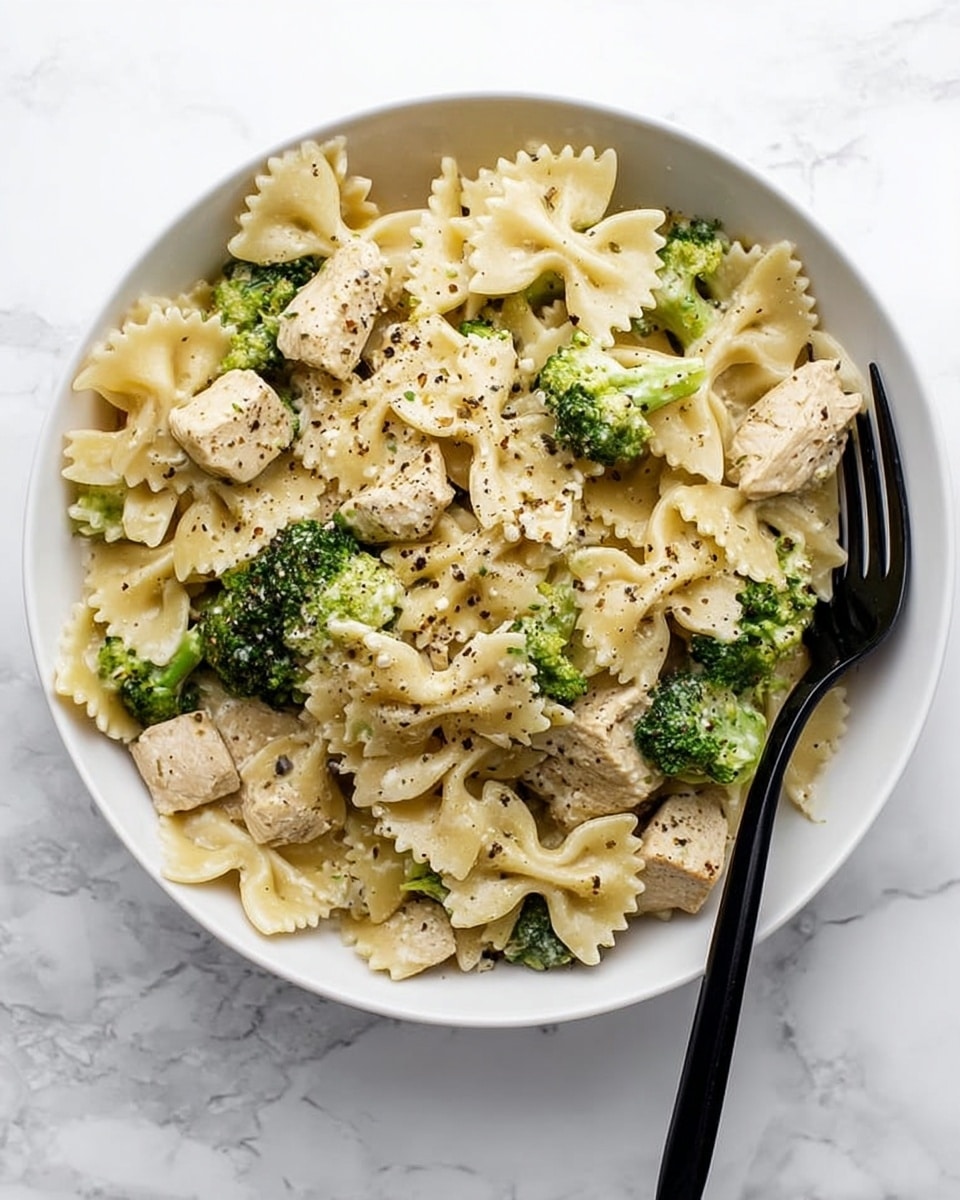 A white bowl filled with creamy farfalle pasta mixed with pieces of cooked chicken and bright green broccoli florets, all coated in a smooth white sauce. The pasta has bow tie shapes with slightly curved edges, evenly spread across the bowl. The chicken pieces are light brown and cubed, nestled among the pasta and broccoli, which add a pop of green. Black pepper is sprinkled on top, adding texture and contrast. On the right side of the bowl, a black fork rests on the edge. The scene is set on a white marbled surface. photo taken with an iphone --ar 4:5 --v 7